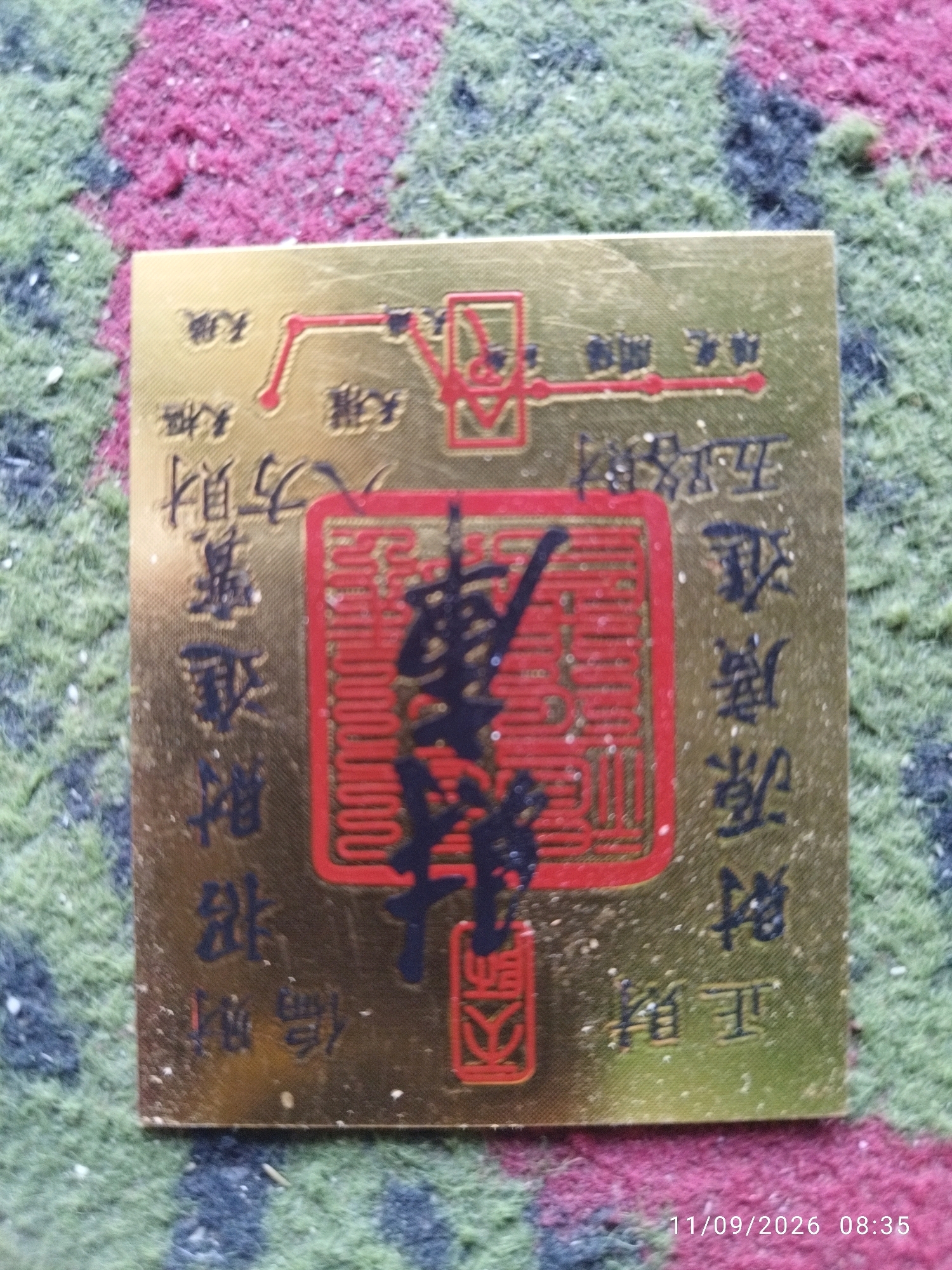 The image shows a close-up of a gold-colored square plaque with red and black text and symbols. The plaque is resting on a textured surface that appears to be a carpet or rug with red, green, and black fibers. The plaque features Chinese characters, some of which are within a red square or rectangular border. The overall impression is of an ornate, possibly decorative or symbolic item. There is a timestamp in the bottom right corner of the image: "11/09/2026 08:35". There are no discernible features in the image that indicate a specific location in the city of Mojokerto, Indonesia.