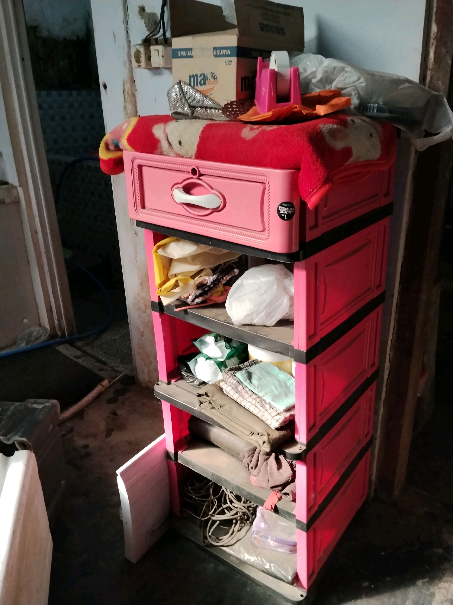 A three-drawer pink plastic cabinet is located inside a dimly lit room, likely in Panggungan, Indonesia. The cabinet is topped with a red blanket featuring a cartoon character, and a silver, sequined clutch bag is placed on the blanket. Various items are stored within the cabinet's open drawers, including white plastic bags, folded fabric, and coiled wires. To the left of the cabinet, a white rectangular object and a ribbed pipe are visible on the floor. The room's walls are dirty and cracked, and an electrical outlet is mounted on the wall above the cabinet. The floor is a mix of concrete and dirt, with some liquid stains.