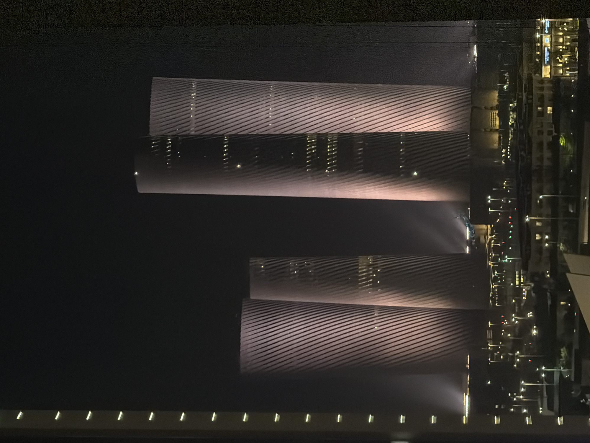 A nighttime exterior shot shows a city skyline at night. Four cylindrical skyscrapers are illuminated with pink light and have a diagonal ribbed pattern on their exterior. The skyscrapers are surrounded by a city with many lights, indicative of an urban environment.