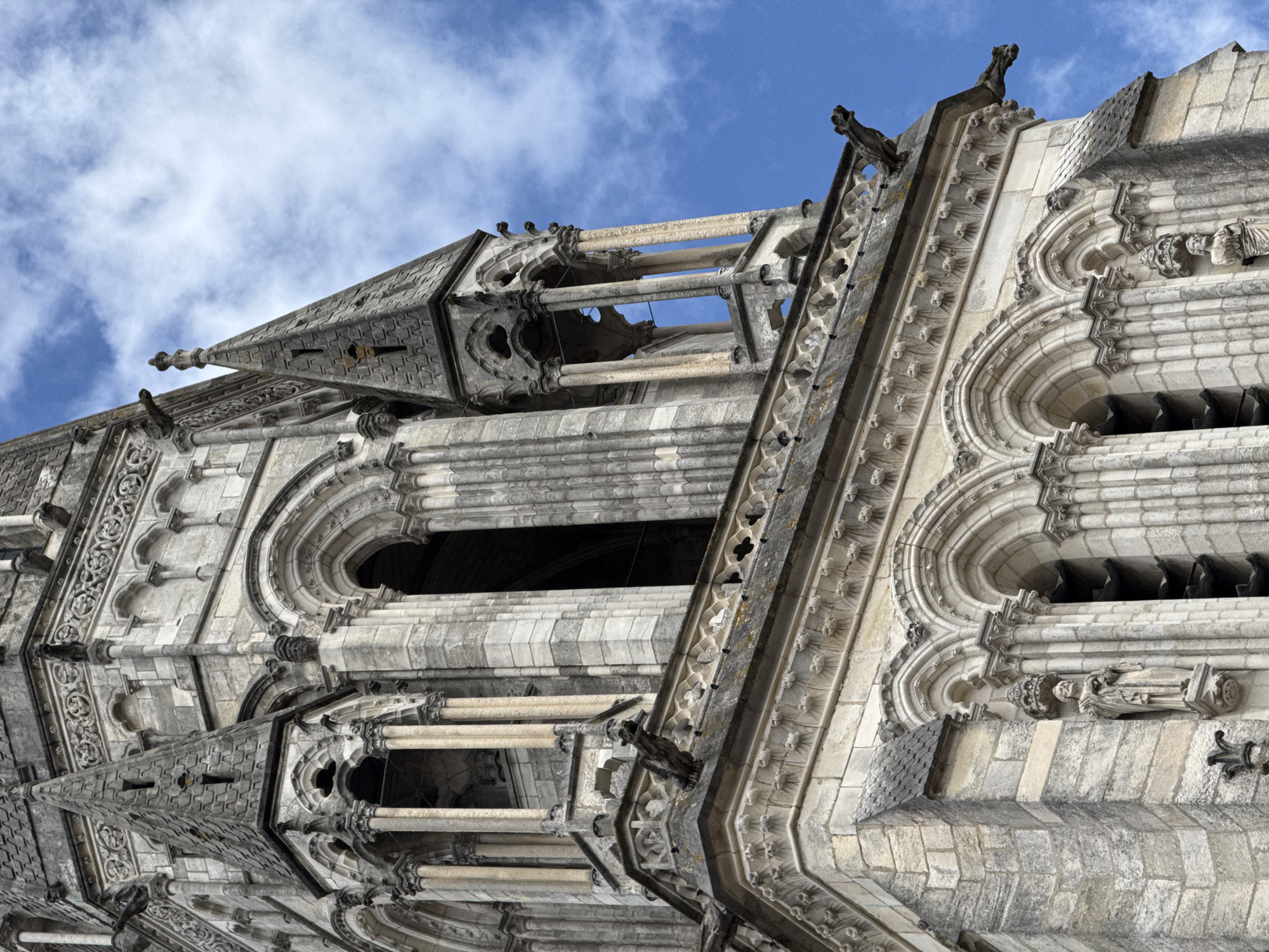 The image shows a close-up, low-angle view of the upper section of the north tower of Nantes Cathedral, also known as the Cathédrale Saint-Pierre-et-Saint-Paul de Nantes, in Nantes, France. The architecture is Gothic, characterized by intricate stone carvings, pointed arches, ribbed vaults, and buttresses. Several gargoyles are visible, perched on the edges of the structure, and small statues are set into niches. The stone is light-colored, weathered, and appears to be in good condition. The sky behind the cathedral is a vibrant blue with scattered white clouds. The image focuses on the detailed stonework and the dramatic upward perspective of the tower against the sky.