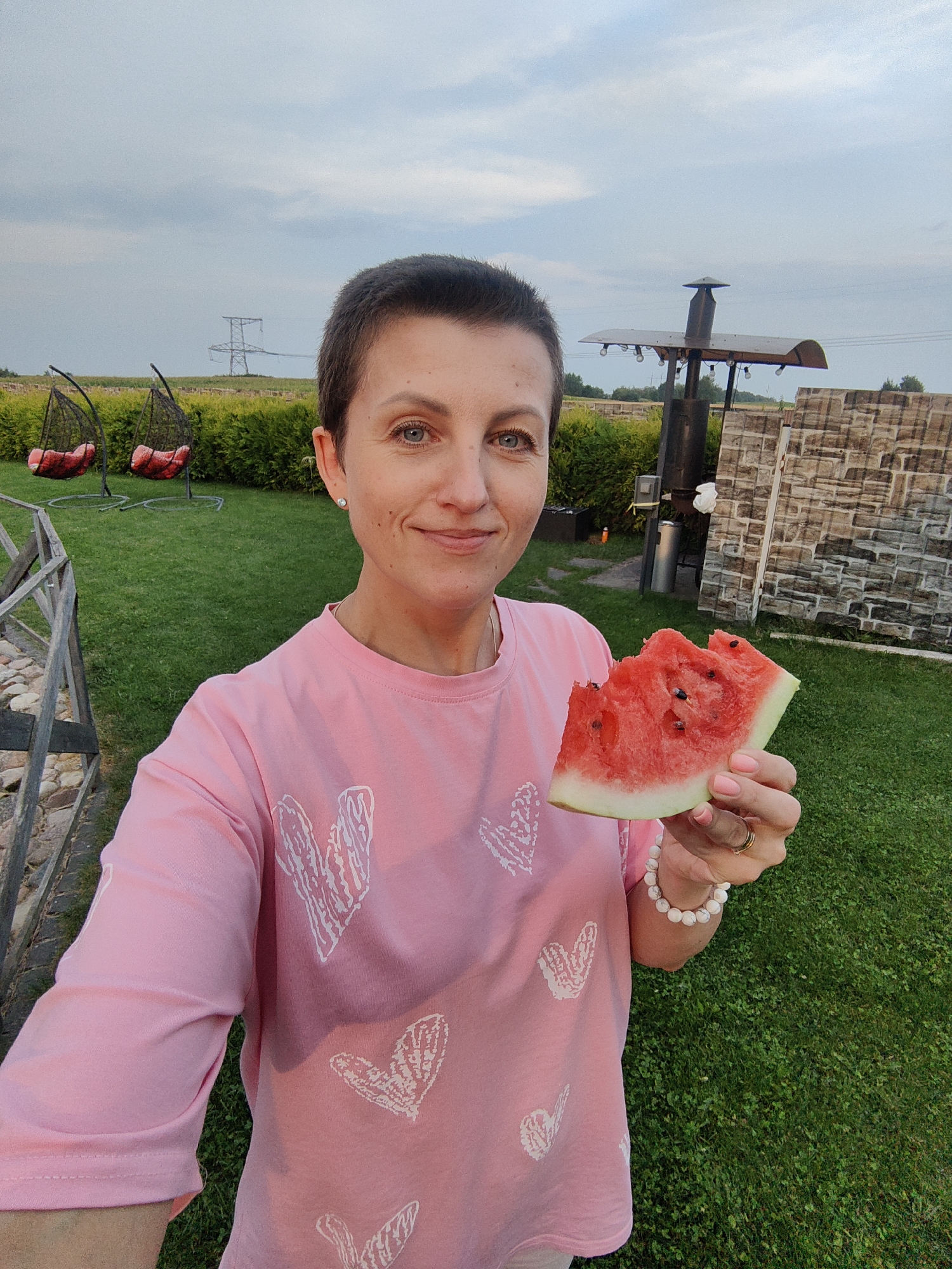 In the city of Фаніпальскі сельскі Савет, Belarus, a woman with short brown hair and blue eyes, wearing a pink t-shirt with white heart patterns and a white beaded bracelet, holds a slice of watermelon in her right hand. She is standing on a green lawn in front of a wooden fence, a hedge, and a stone wall. In the background, two hanging chairs with red cushions are visible. The sky is partly cloudy.