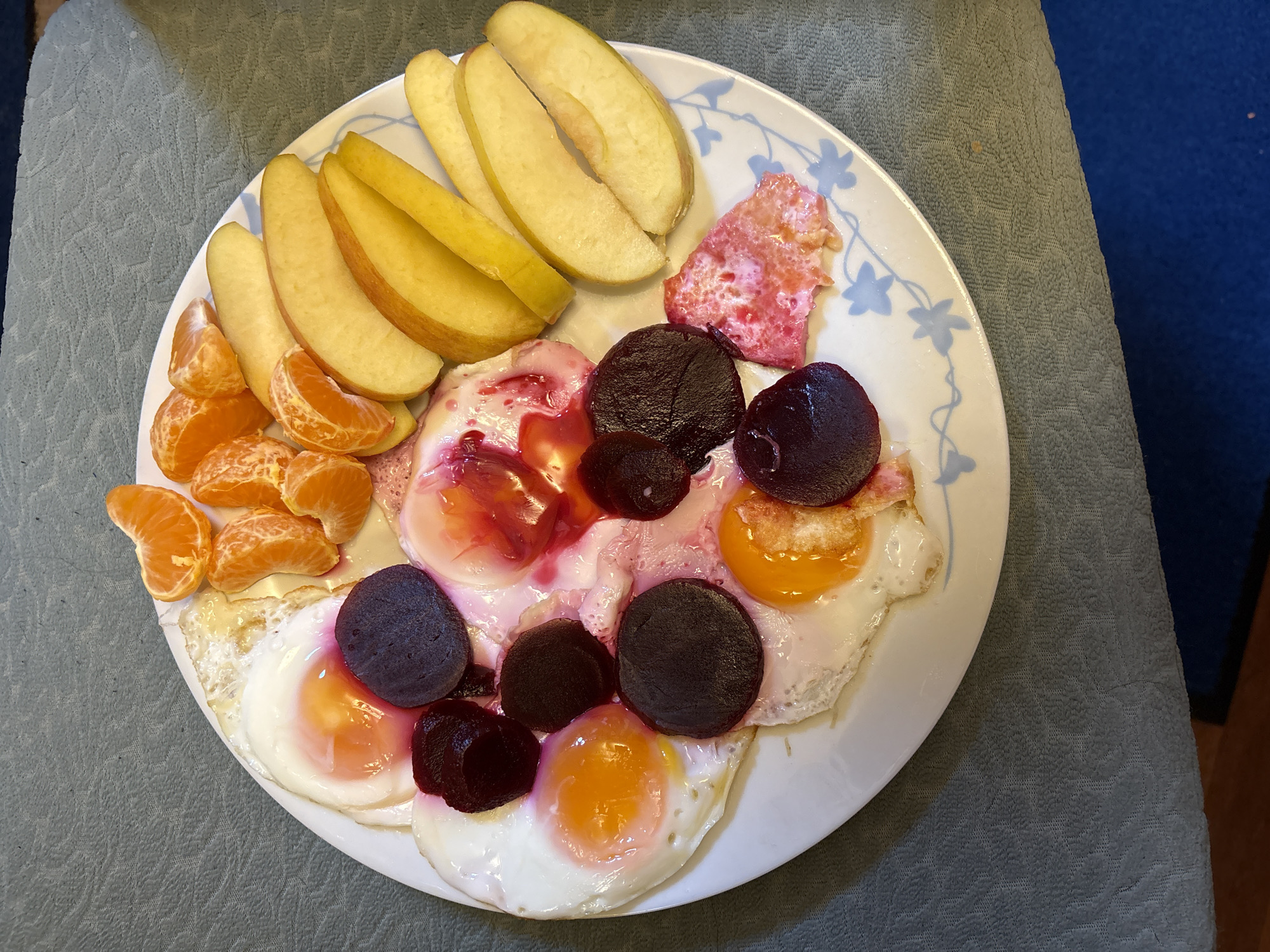 A plate of food is arranged on a light gray, textured surface, likely a tablecloth or placemat. The plate appears to be white with a blue floral pattern around the edge. The food items include: sliced apples (yellowish color), mandarin orange segments, fried eggs (several), sliced beets (dark purple), and a piece of cooked meat or other protein (pinkish-red). The eggs have been topped with the sliced beets and the pinkish-red meat. The scene does not provide sufficient information to verify the location as Oswestry, United Kingdom.