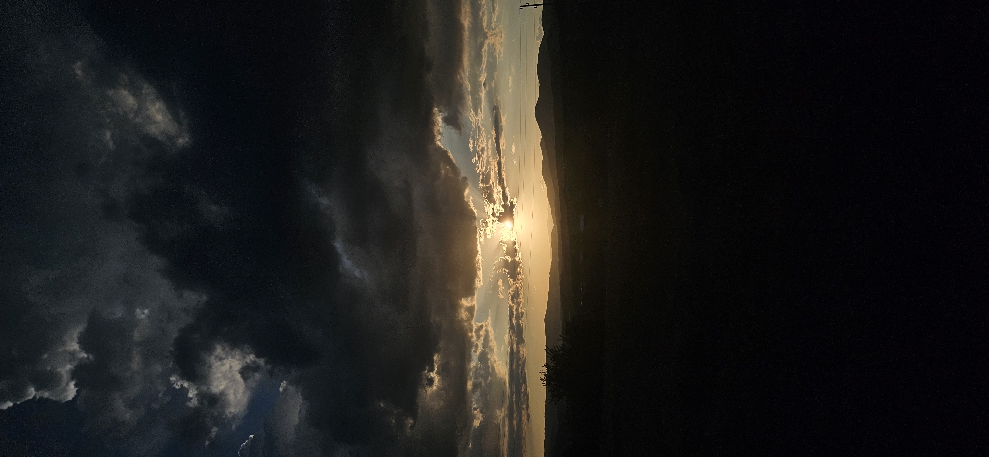The image shows a horizontal view of a sunset. The left side of the image features dark storm clouds, while the right side is in darkness. In the center, the sun shines brightly between the clouds, creating a vivid golden light. Silhouetted mountains are visible on the horizon behind the sunlight. Power lines are present on the right edge of the lighted portion. The scene is likely located near the city of Momin Prohod, Bulgaria, as indicated by the tags. The image captures a moment of contrasting light and shadow during a sunset, emphasizing the interplay of the sun, clouds, and landscape.
