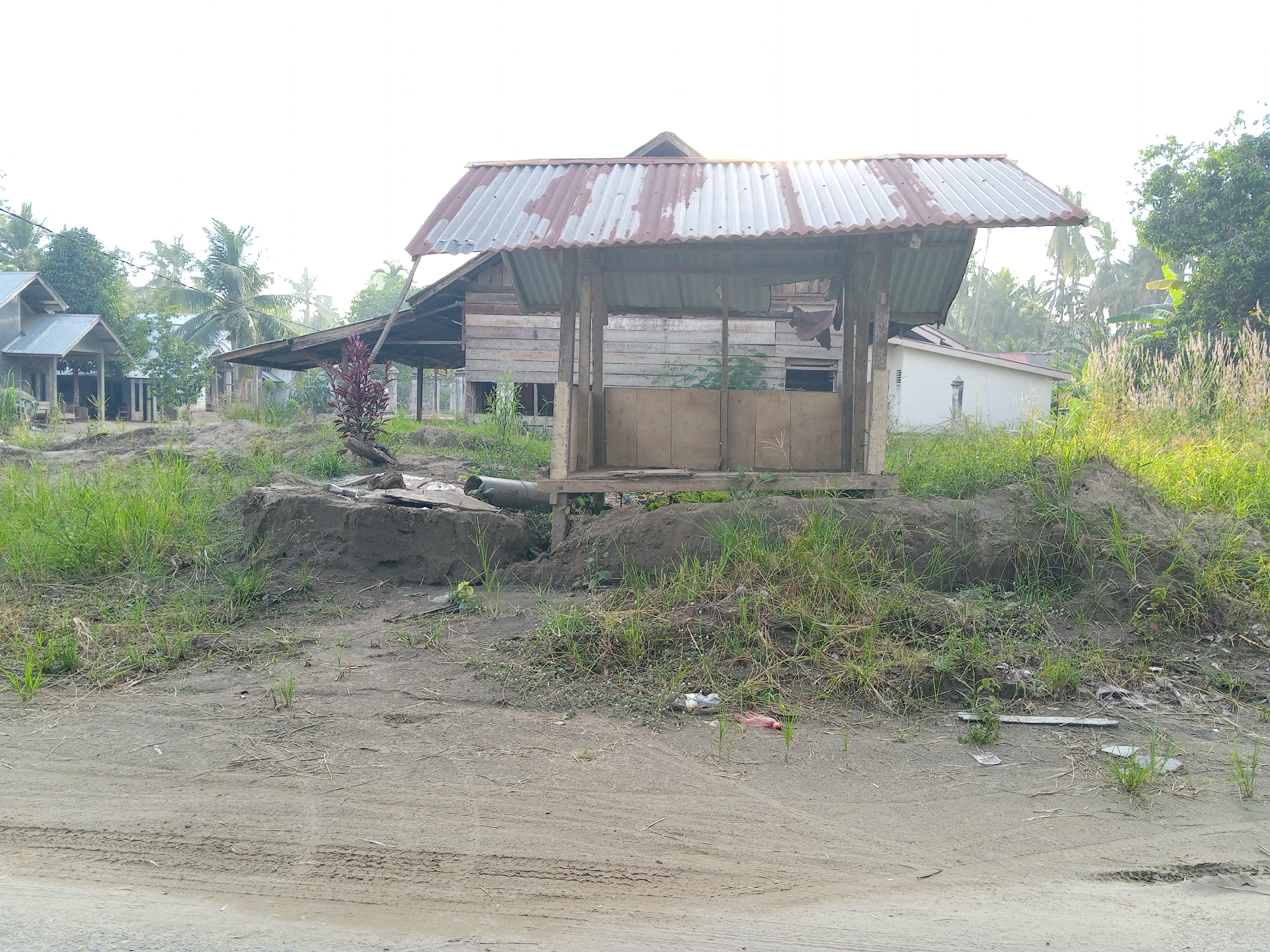 The image depicts a small, dilapidated wooden structure with a corrugated metal roof, situated in what appears to be a rural or semi-rural area near Kruengbeukah, Indonesia. The structure is elevated on posts and has a partially enclosed front. The ground is sandy and covered with patches of grass and weeds. There is visible litter scattered in the foreground. Other wooden buildings and vegetation, including palm trees, are present in the background, suggesting a residential or agricultural setting. The lighting indicates it is daytime, possibly early morning or late afternoon. Tire tracks are visible on the dirt road in the foreground.