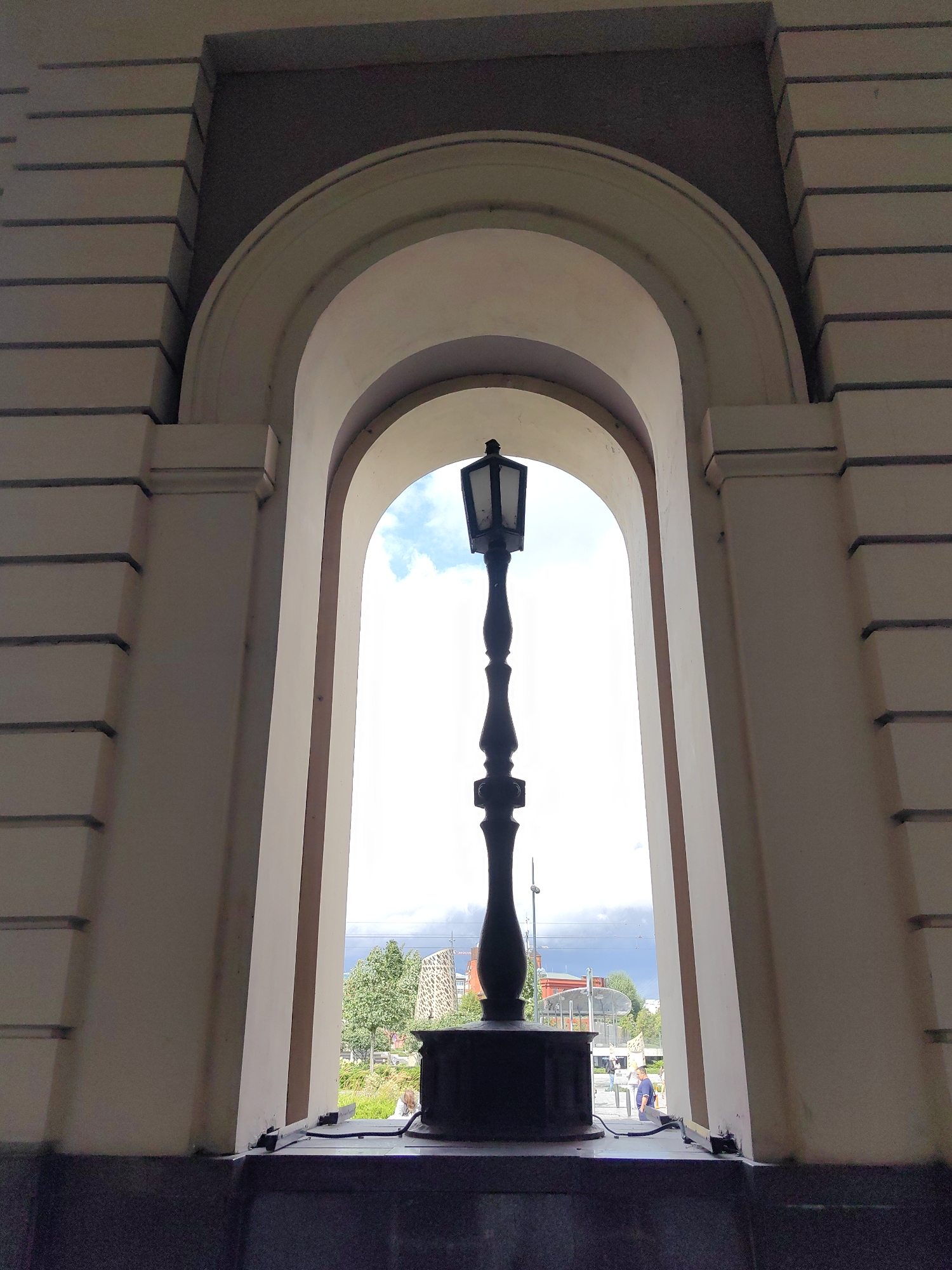 A dark, ornate lamppost stands silhouetted against a bright sky, framed by a large architectural archway. The archway is part of a building with textured cream-colored walls featuring horizontal indentations. Through the arch, a park-like area is visible, with trees, some modern buildings, and a few people walking in the distance. The sky is partly cloudy, with a darker cloud formation on the horizon. The ground in the foreground appears to be dark stone.