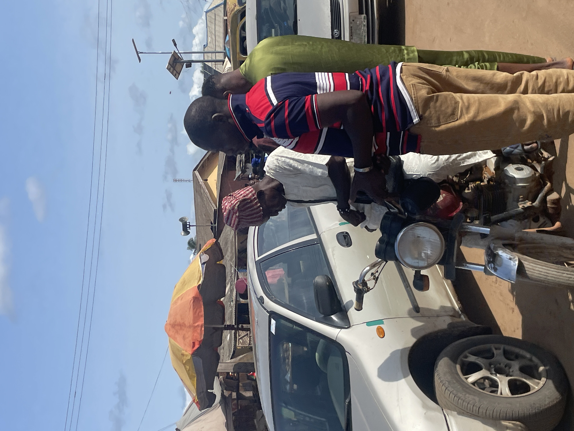 The image shows three men near a white car and a motorcycle in Ajibo, Nigeria. One man, dressed in a red and blue striped shirt and khaki pants, appears to be working on the motorcycle. Another man, in a white shirt and red patterned head covering, is also focused on the motorcycle. A third man, wearing a green shirt and khaki pants, stands behind them. A multi-colored umbrella is visible nearby, along with utility poles and power lines against a blue sky. Other vehicles and buildings are visible in the background.