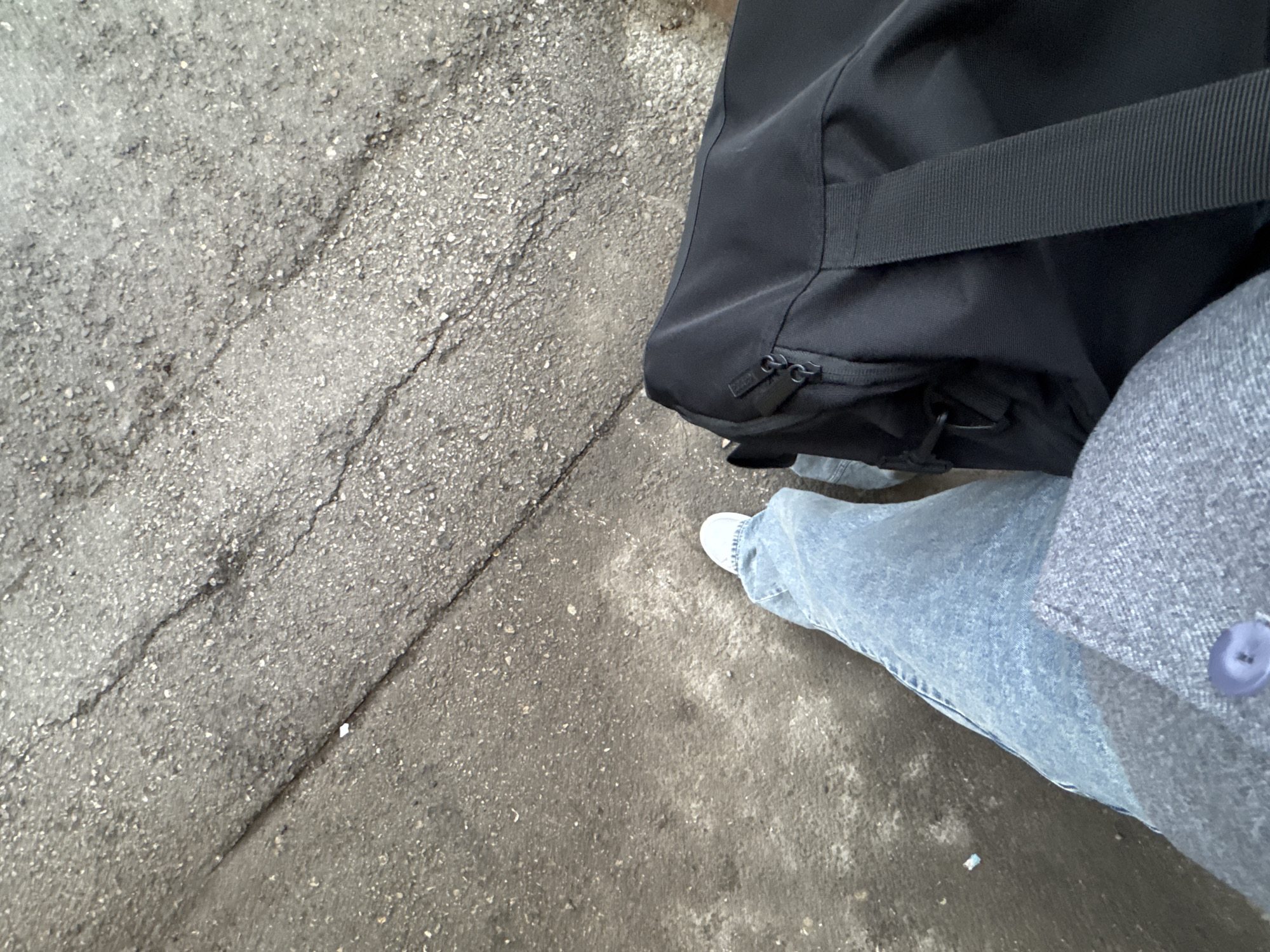 The image shows a view from above of a section of cracked concrete sidewalk in Perm, Russia. A portion of a black duffel bag occupies the upper right quadrant. A person wearing light blue jeans, white sneakers, and a gray jacket with a purple button is standing beside the bag. No actions or interactions are visible.