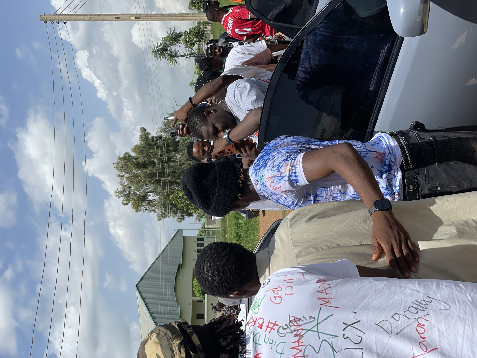 The image depicts a group of people gathered around a car in what appears to be an outdoor setting in Durumi, Nigeria. The sky is partly cloudy, suggesting it is daytime. Several individuals are holding up their phones, seemingly capturing photos or videos of something or someone within the group or near the car.
One person in the foreground, wearing a white t-shirt with various handwritten messages and drawings in different colors, is prominently featured. Another person in the mid-ground, wearing a blue and white patterned shirt, is also visible. A man in a red jersey with the name "OGUNDIMUTI" and the number "9" on the back is seen among the crowd, leaning into the car. A person wearing a black beanie and a black jacket is also present. The scene suggests a gathering, possibly related to an event or a celebrity appearance, given the attention from the onlookers with their phones.
The overall mood seems to be one of engagement and excitement, with people focused on their devices and interacting with each other. The presence of a utility pole and power lines, along with trees and a building with a metal roof in the background, provides environmental context. The activity appears to be a spontaneous or informal gathering where people are documenting the moment.