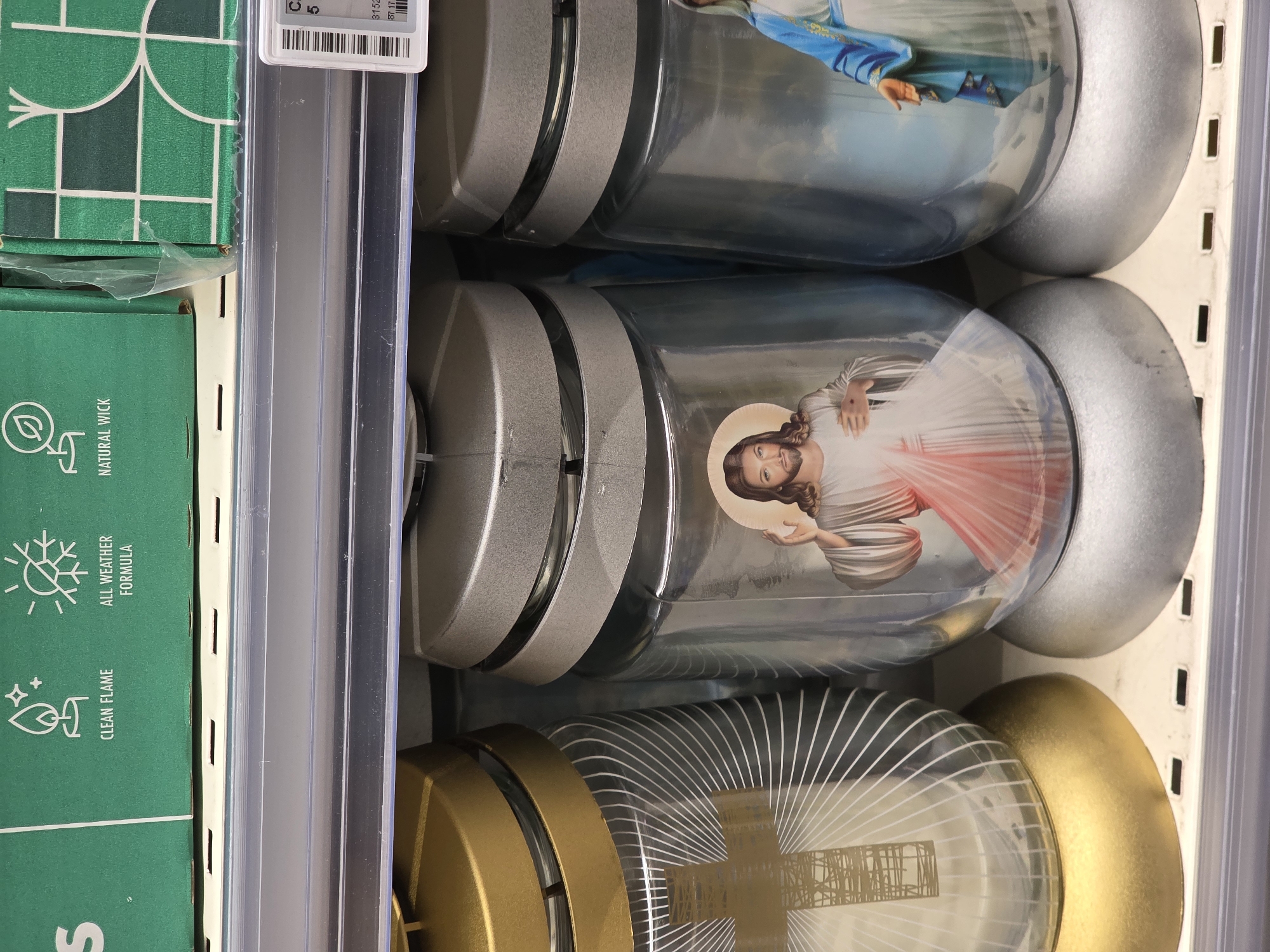 The image shows a shelf in a store displaying several memorial candles. The candles are glass jars with metallic lids, some gold and some silver. Two of the candles prominently feature religious imagery. The candle in the center has an image of Jesus Christ with outstretched arms and a halo. To its right, another candle shows a figure, likely a saint or the Virgin Mary, in a blue robe. To the left, a candle has a stylized cross design. Above the candles, a green cardboard box with text and icons related to candles is partially visible, indicating product information such as "CLEAN FLAME," "ALL WEATHER FORMULA," and "NATURAL WICK." A price tag with a barcode is attached to the shelf in the upper right corner. The shelf appears to be in a retail environment, possibly a supermarket or a specialized store. The context of Bucharest, Romania, is not directly evident from the visual information in the image itself.