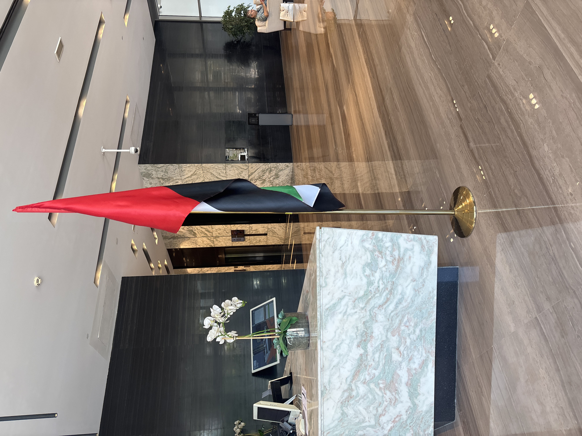 The image depicts the interior of a modern lobby, likely in Dubai, United Arab Emirates, given the flag displayed. A prominent United Arab Emirates flag stands on a golden pole in the foreground. To the left, a reception desk features a marble countertop with an orchid plant and electronic devices. Behind the desk, a dark, textured wall serves as a backdrop. In the mid-ground, a person is seated on a sofa to the right, engaged with a mobile device, with shopping bags placed nearby. The floor is highly polished wood or marble, reflecting the ambient lighting and architectural details. The ceiling features recessed lighting and a security camera. The overall atmosphere is one of a clean, upscale commercial or hospitality space.