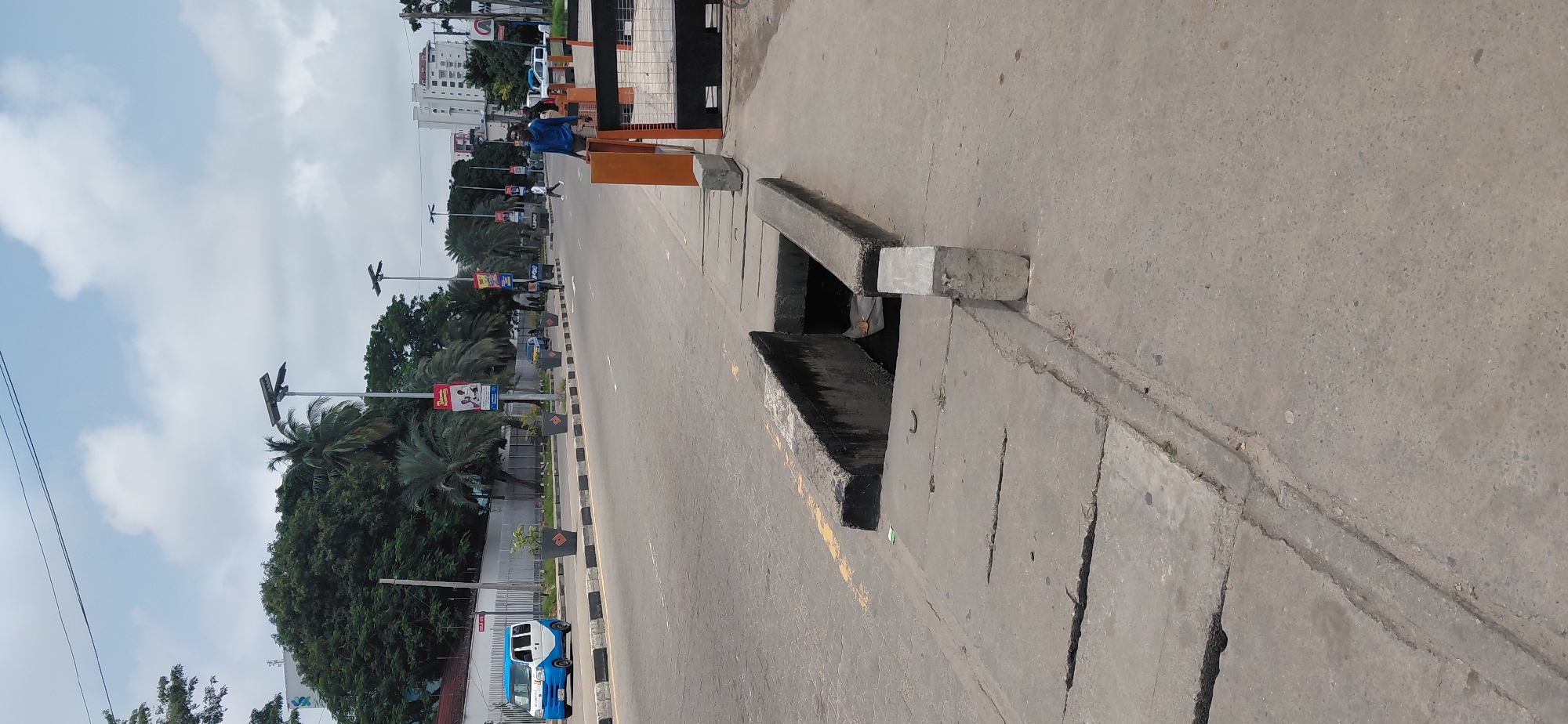 The image depicts a street scene in the city of Itirin, Nigeria. In the foreground, a large, open concrete drain runs along the side of the road, with a concrete cover partially in place. The road itself is asphalt with yellow and black painted borders. Several planters with small trees are placed along the median of the road, which is bordered by a black and white striped curb.
In the middle ground, a light blue and white minivan is driving on the road. Further down the road, several pedestrians can be seen walking. There are also tall streetlights with solar panels visible along the road. In the background, there are trees, some buildings, and a bright sky with scattered clouds. A man in a blue shirt is standing near an orange and black barrier on the sidewalk.