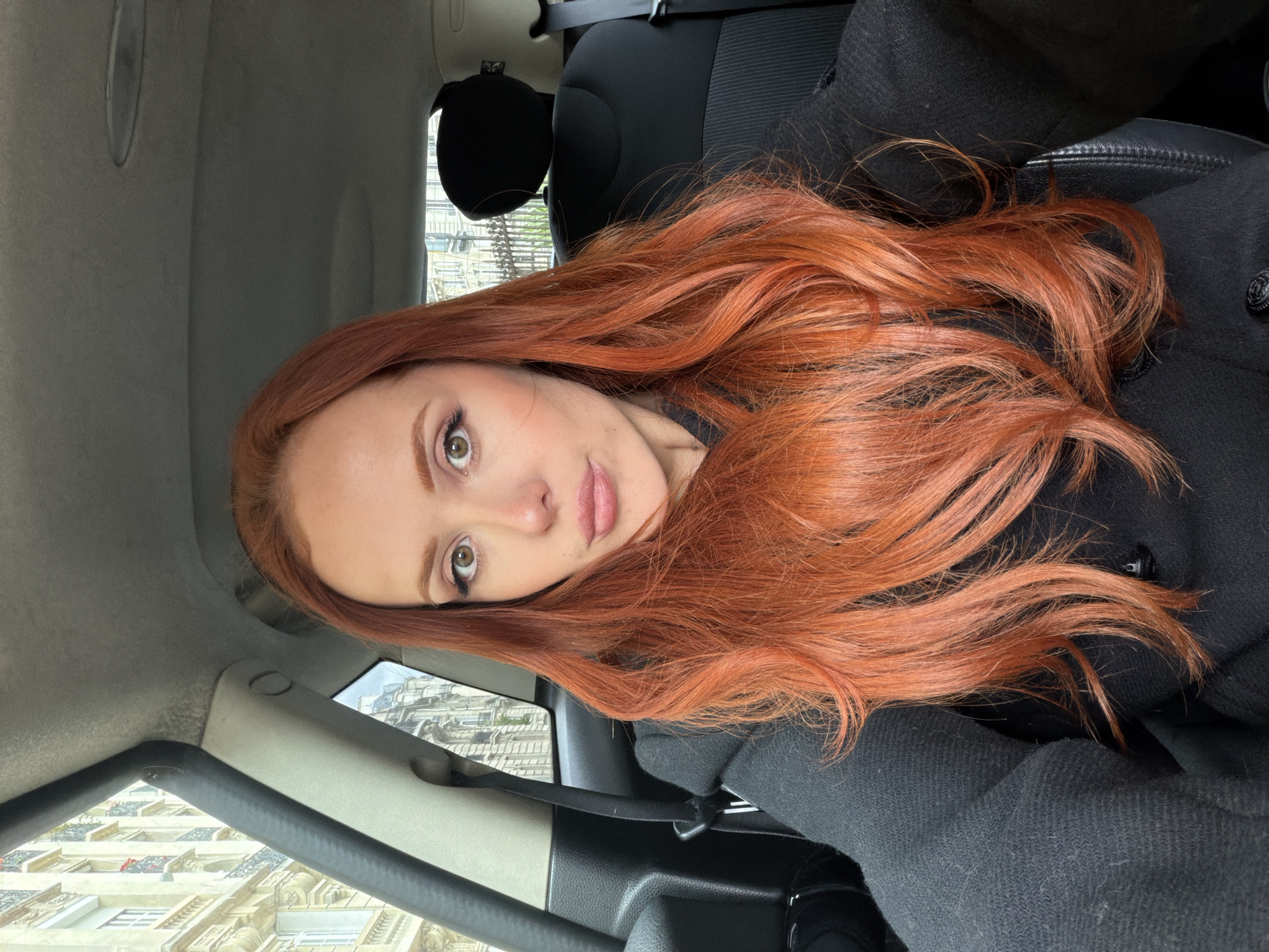 A woman with long, red, wavy hair is seated in the driver's seat of a car. She is wearing a black coat. Outside the car window are buildings characteristic of Paris, France. The architecture includes off-white facades with ornate details and balconies, suggesting a location within the city of Paris. The photo appears to have been taken inside a parked car, likely during the daytime given the natural light.