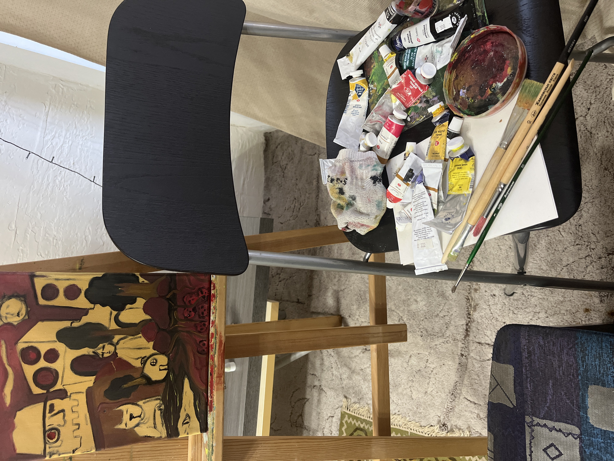 The image displays an active art workspace, likely indoors, captured from an overhead angle. On the left, a partially visible abstract or semi-abstract painting rests on a wooden easel, characterized by bold brushstrokes and a palette of red, gold, and black, depicting stylized figures or faces.
To the right, a black chair or stool serves as a makeshift table, cluttered with various art supplies. Numerous tubes of paint are scattered across its surface, including brands like "Sonnet" and labels indicating colors such as "Cadmium lemon (HUE)," "ЖЕЛТЫЙ" (Yellow), "КРАСНЫЙ" (Red), and "КОБАЛЬТ" (Cobalt), with some specifying "АКРИЛОВАЯ" (Acrylic). A well-used palette, containing mixed red and brown paint, sits among the tubes. Several paintbrushes are also present, some marked "Живописные кисти" (Painting brushes) and "ЩЕТИНА" (Bristle), with one noted as "Художник Россия Киров" (Artist Russia Kirov). A paint-splattered white rag lies nearby.
The background features a light-colored, textured rug or carpet on the floor, and a white, textured wall with a string of small lights. Part of another chair, upholstered in blue fabric with a geometric pattern, is visible in the bottom right corner. The scene clearly indicates ongoing artistic activity within a creative and lived-in environment.