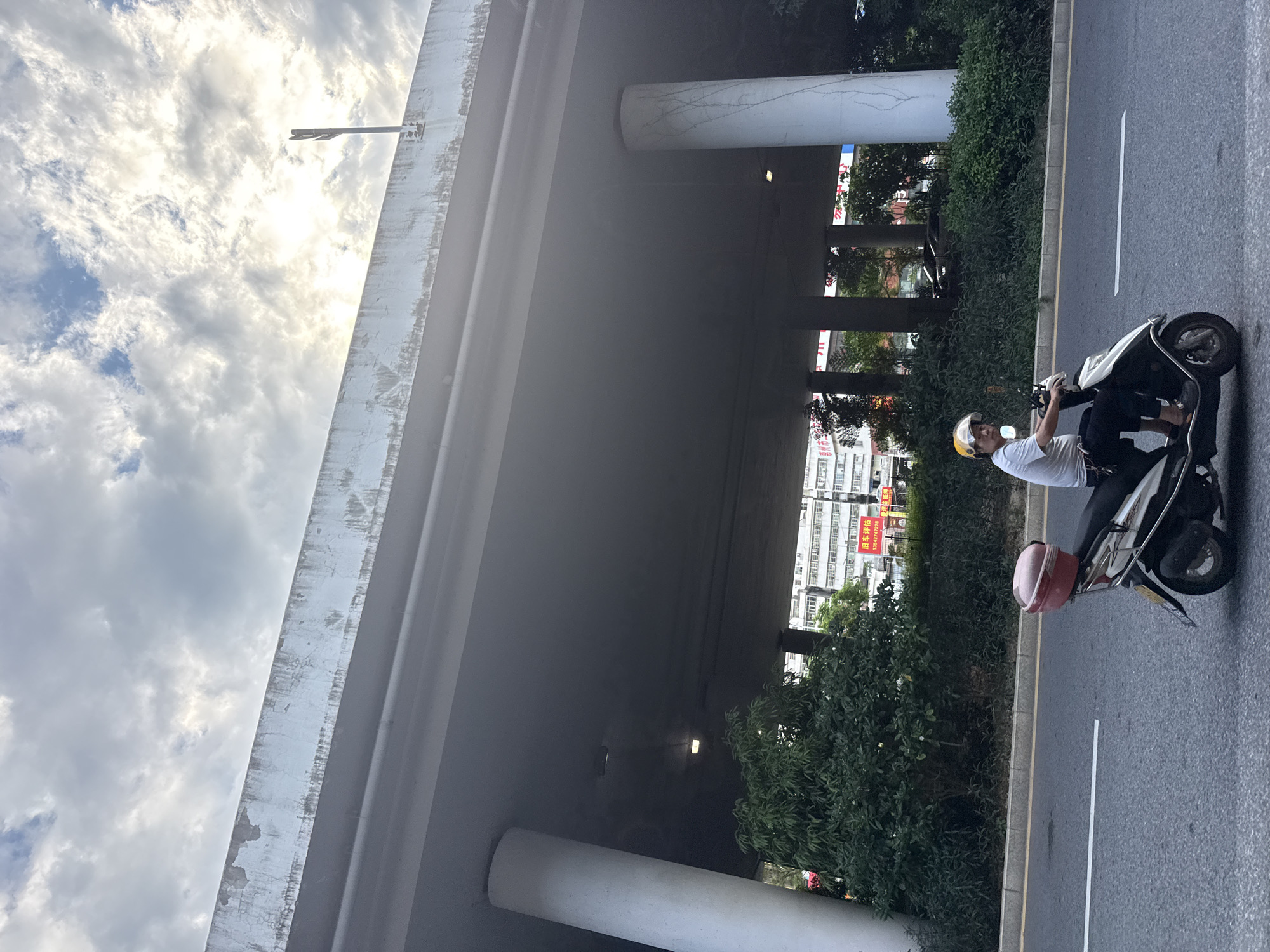 A person on a scooter rides on a paved road in Huizhou, China, beneath an elevated highway. The person, wearing a white t-shirt and a yellow helmet, is positioned on the right side of the scooter, which has a red storage box on the back. The elevated highway is supported by large concrete pillars. Green trees and bushes line the side of the road. In the background, buildings and signage are visible. The sky is cloudy, with sunlight filtering through.