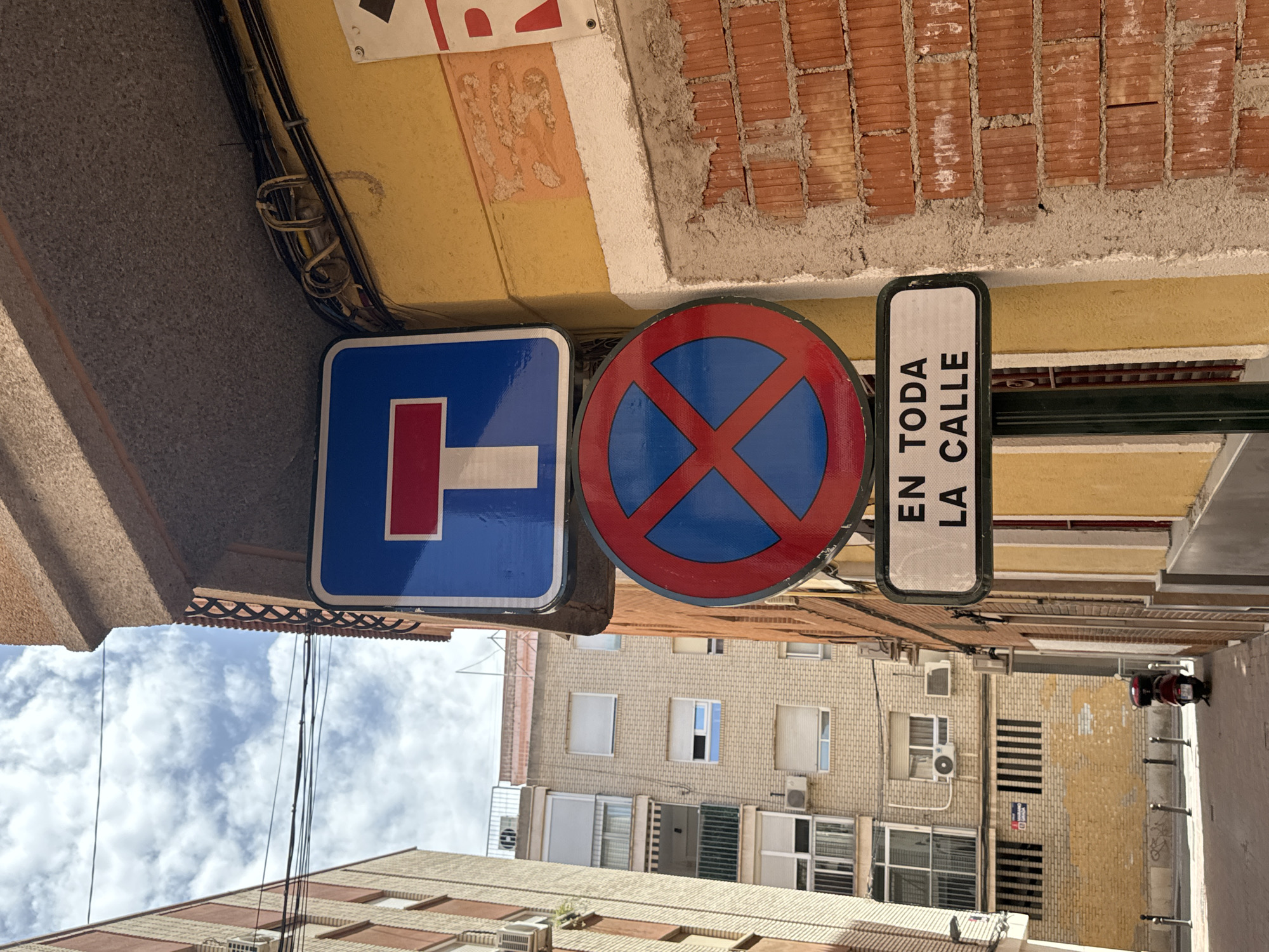 The image shows a street in the city of Murcia, Spain, with three signs mounted on a pole. The topmost sign is a blue square with a white horizontal bar and a red vertical bar forming a "T" shape, indicating a dead end. Below it is a circular red and blue sign with a red "X" inside, signifying no stopping or parking. The bottom sign is a rectangular white plate with black text that reads "EN TODA LA CALLE," which translates to "in the entire street."
The signs are attached to a pole which is positioned against a yellow building. To the left of the pole, a multi-story residential building with light-colored brickwork and numerous windows is visible. Further to the left, another building with orange brickwork and several air conditioning units can be seen. The street is paved and has several bollards lining the side. In the background, a red scooter is parked. The sky is cloudy, with patches of blue. The scene suggests a residential area within a Spanish city.