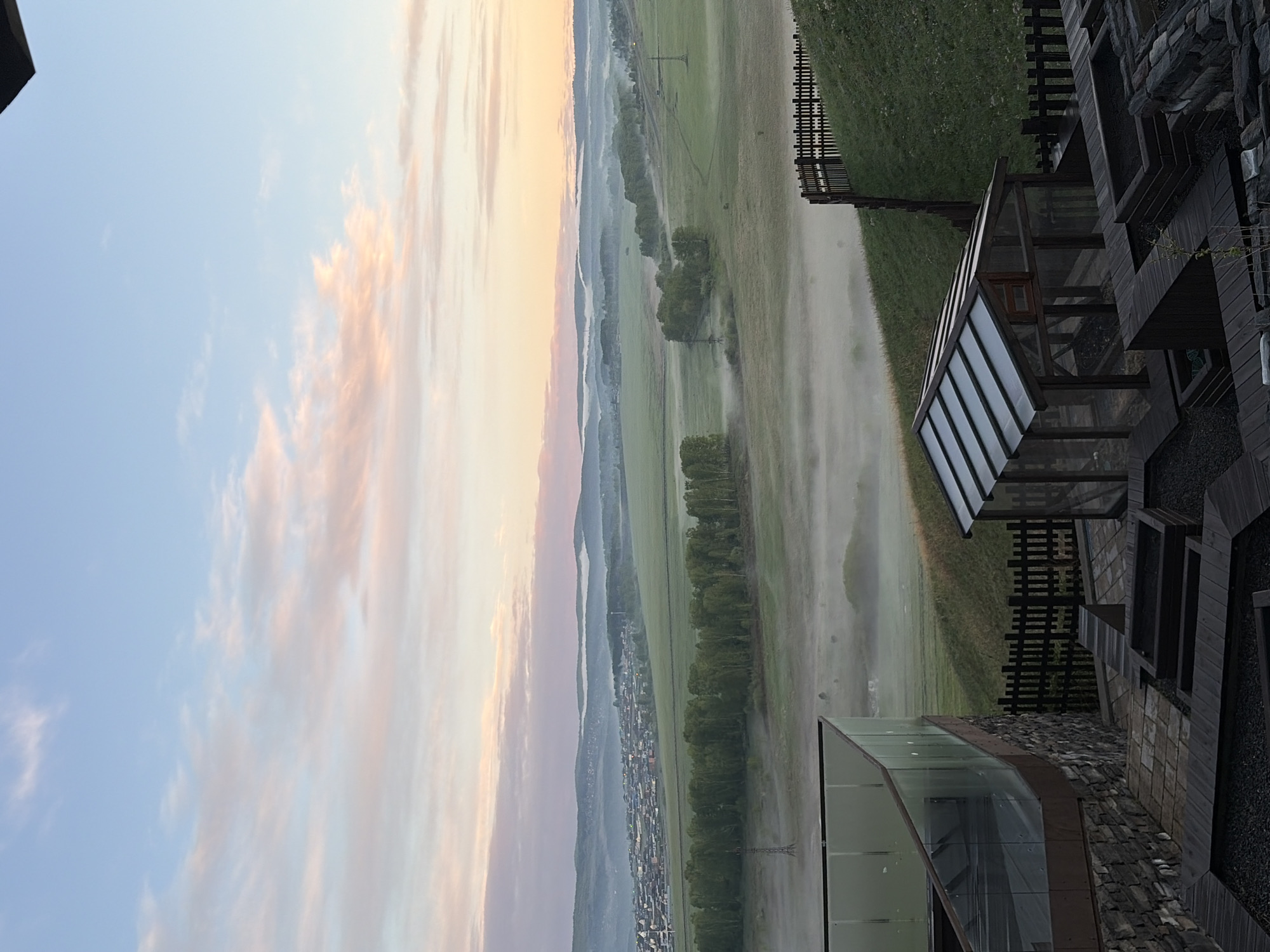The image shows a landscape viewed from a building, potentially in or near a town. The scene includes a wide expanse of green fields partially obscured by fog or mist. A line of trees runs along a portion of the fields, and beyond that, there is a view of a town nestled in a valley or at the foot of hills or mountains. In the sky, there's a mix of blue and pastel colors, suggestive of either dawn or dusk. Parts of the building from which the image is taken are visible in the frame, showing a modern architectural style with glass and dark wood elements. The architectural details include a glass-covered patio or balcony area. The fence runs along a portion of the fields near to the building. There is no evidence to confirm if this is located in Shamoni, Russia.