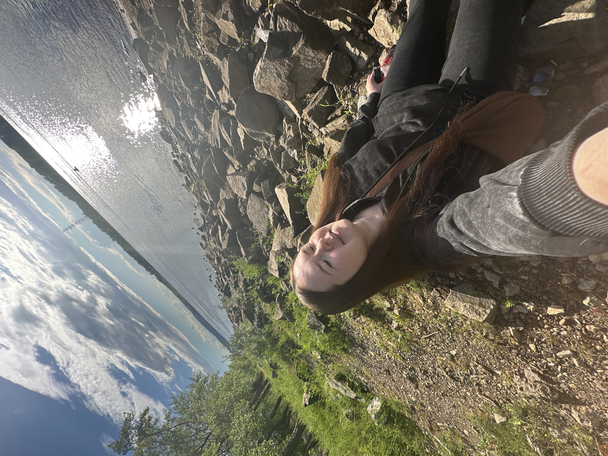 A young woman lies on a rocky shore next to a body of water, with trees and greenery visible on the bank. She is wearing a dark gray hoodie and black pants. She is taking a selfie, smiling at the camera. The sun is shining brightly, reflecting off the water. The sky is blue with some scattered white clouds. The scene appears to be set during the daytime, with bright sunlight indicating a sunny day. The rocky terrain and the presence of water suggest an outdoor natural environment, possibly a lake or riverbank. The woman's relaxed posture and smile convey a sense of happiness and enjoyment of her surroundings. There is no visible text in the image.