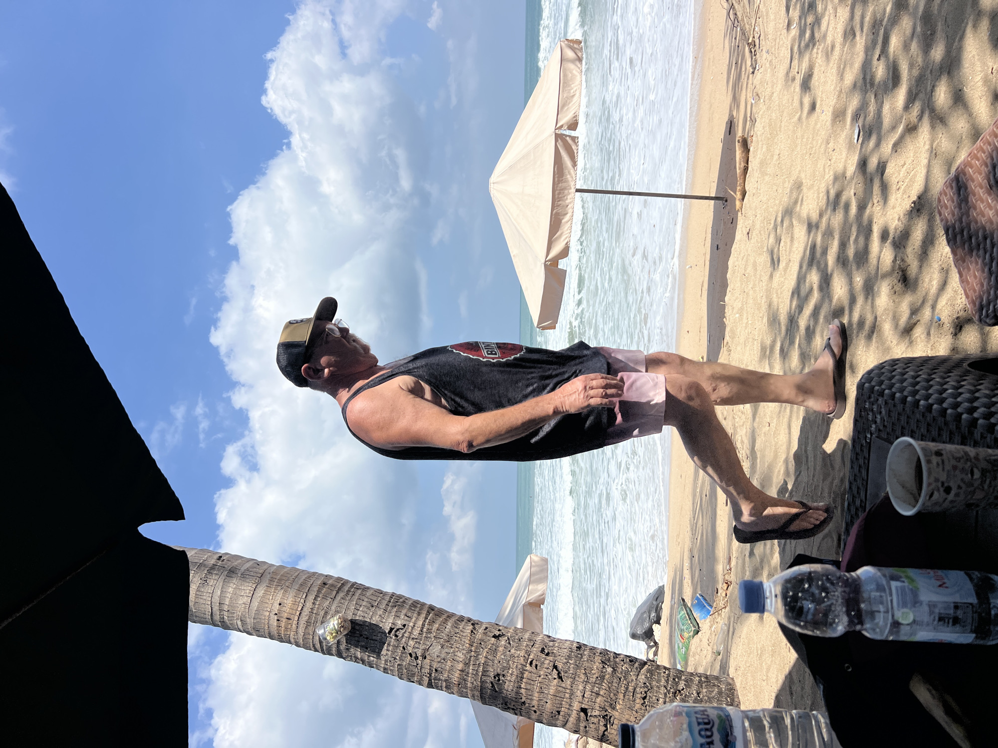 A man with glasses and a baseball cap walks along a sandy beach in Kuta, Indonesia. He is wearing a dark tank top and light-colored shorts. The ocean is visible behind him with waves rolling onto the shore, and a beige beach umbrella is set up on the sand. In the foreground, there are two bottles of water and a cup on a woven table, along with a portion of a black umbrella and a palm tree trunk. The sky is blue with scattered clouds.