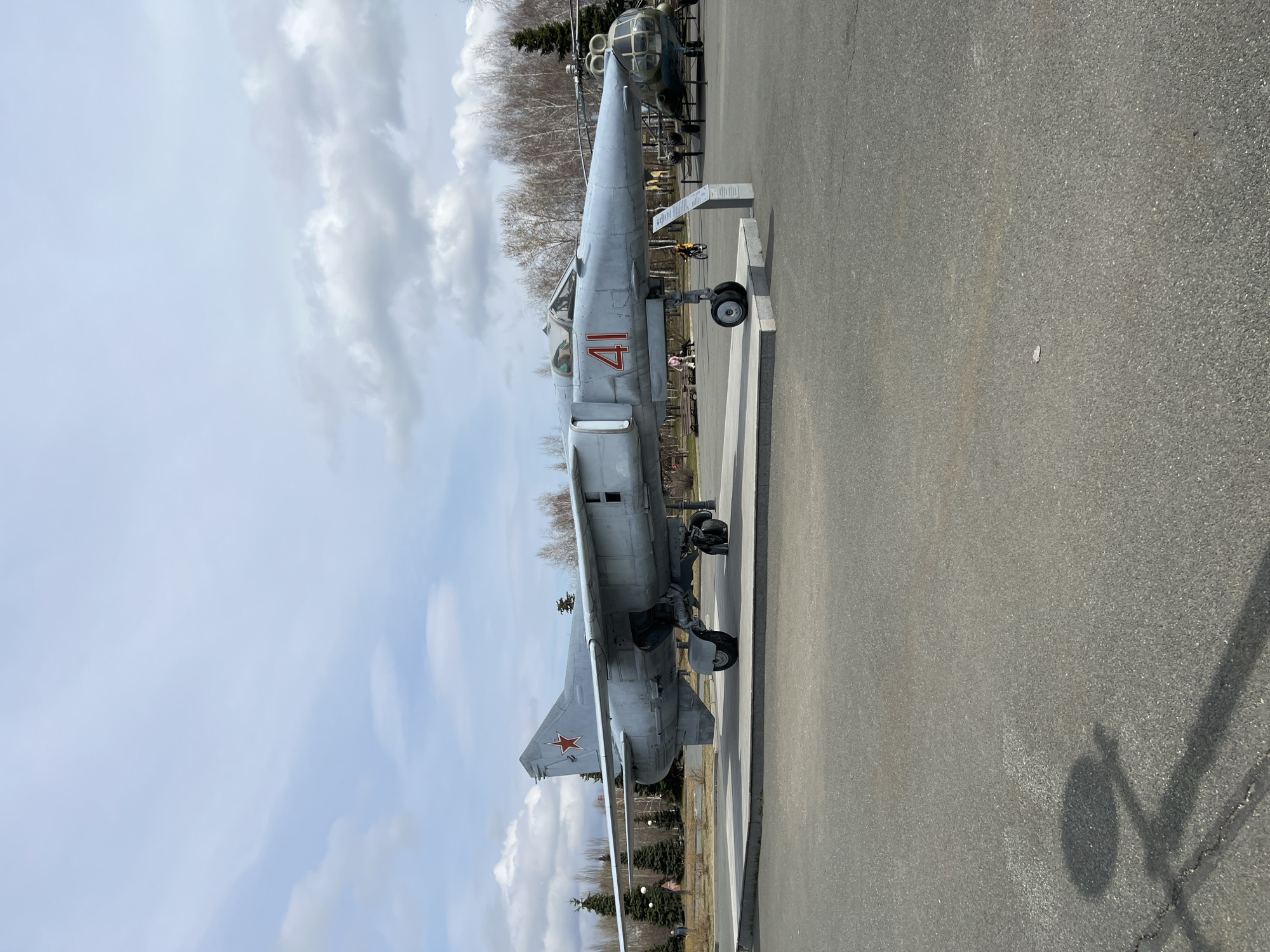 The image displays a grey military fighter jet, likely a Soviet-era model such as a MiG-23 or similar, exhibited outdoors. The aircraft is oriented with its nose pointing towards the right side of the frame and its tail towards the left. A prominent red number "41" is painted on the side of its fuselage, just below the cockpit. A distinctive red star emblem, characteristic of Soviet and Russian military aviation, is visible on the vertical tail fin.
The fighter jet is positioned on a paved asphalt surface, suggesting it is a static exhibit in a museum, park, or public display area. In the background, bare deciduous trees indicate the season is either early spring or late autumn. Another aircraft, possibly a helicopter or a different fixed-wing aircraft, is partially visible behind the nose of the main jet, further reinforcing the impression of an aviation exhibition. The sky above is partly cloudy, with scattered white clouds against a light blue background. Bright daylight illuminates the scene, casting shadows onto the asphalt. No people or active events are discernible in the image, focusing solely on the static display of military hardware. The setting is consistent with a public display in Kazan, Russia.