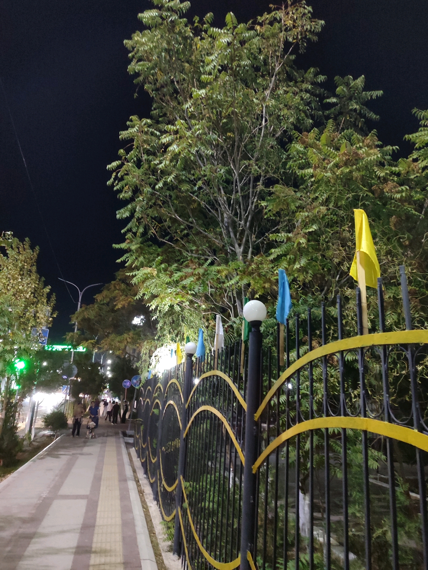 The image depicts a nighttime scene in Nukus, Uzbekistan, featuring a paved pedestrian walkway alongside a black metal fence adorned with yellow decorative elements. Several trees with green foliage are visible, some illuminated by artificial lights. Small flags, including a yellow one and a blue one, are attached to poles along the fence. A lamppost with a spherical light fixture is also present. In the background, several individuals are walking along the path, some accompanied by a dog. The overall environment suggests a well-lit public area, possibly a park or a street lined with greenery.
