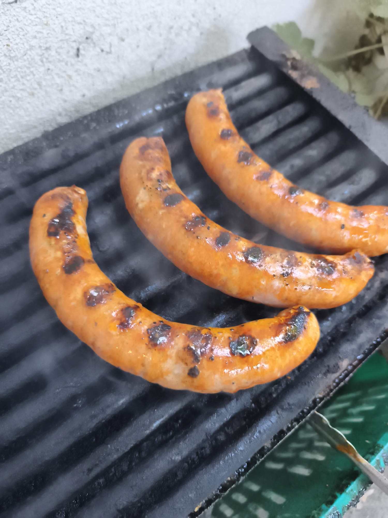 Three sausages are grilling on a black metal grate, some of them are slightly charred. The grate is placed on a concrete surface, possibly a patio or outdoor kitchen area. In the background, there is a hint of a leafy green plant and a textured white wall. A green bin and a pair of tongs are visible in the lower right corner. The scene suggests a casual outdoor cooking setting in Plovdiv, Bulgaria.