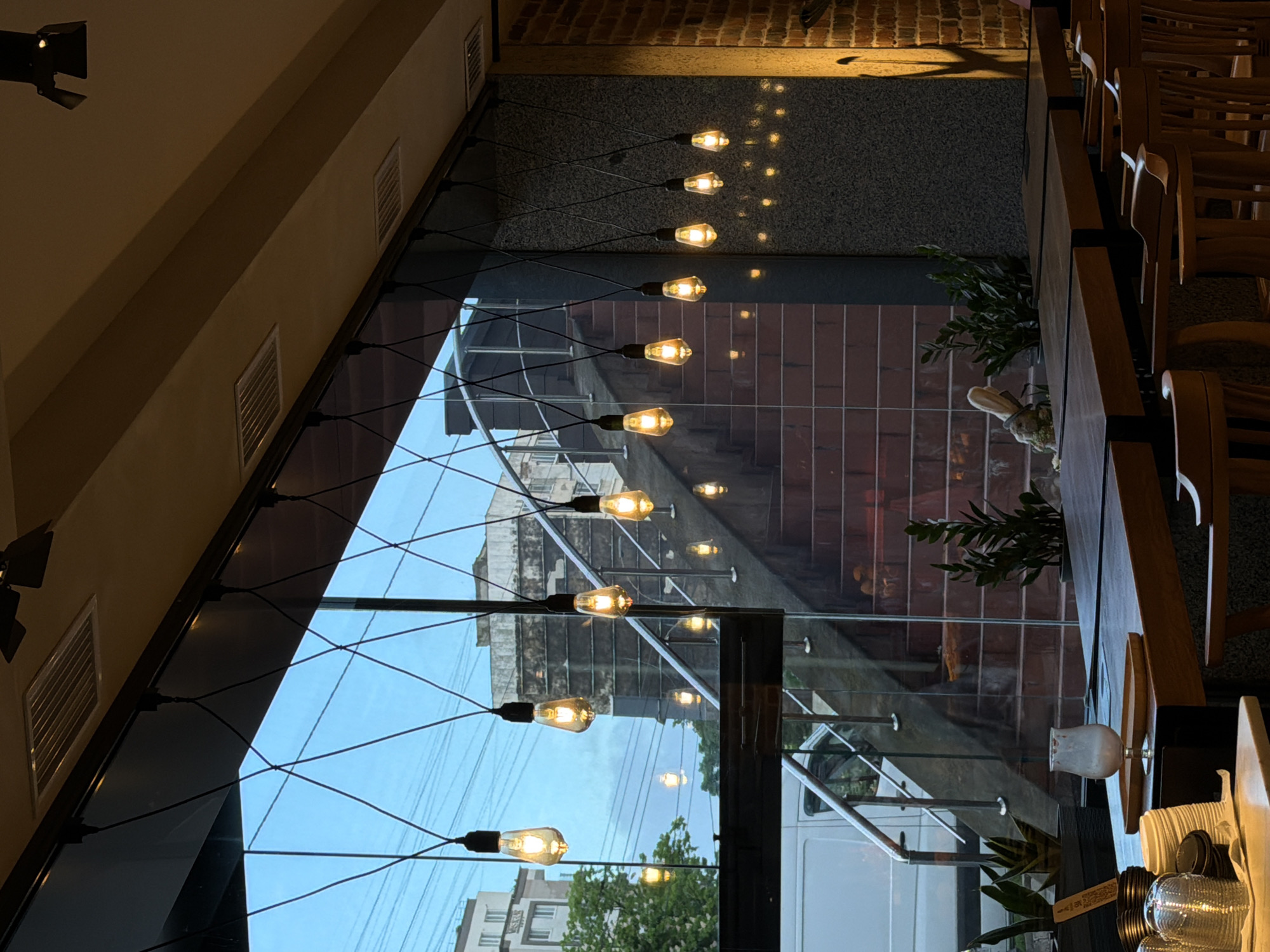 The interior view shows a restaurant setting. The perspective is looking out through a large window, angled and lined with multiple hanging vintage-style light bulbs. The outside view through the window includes a residential area with apartment buildings and trees, with an overcast sky. Inside, adjacent to the window, is a dining area with wooden chairs and tables. A small potted plant with greenery sits on one of the tables, along with a vase and some tableware. The overall ambiance suggests a modern urban eatery with a view.