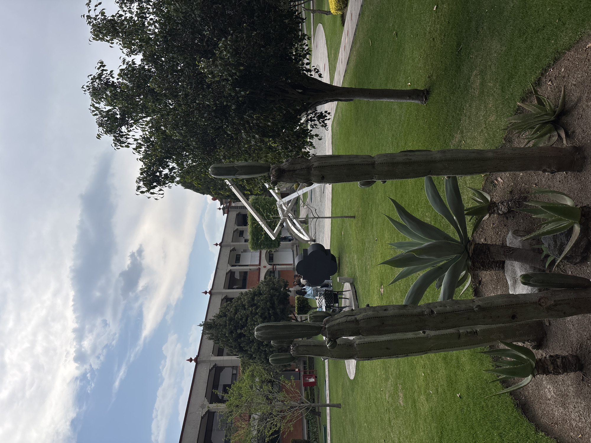 The image depicts a garden scene in San Andrés Cholula, Mexico. A well-manicured lawn is bordered by several tall cacti and an agave plant in the foreground. In the background, a building with multiple floors is visible, along with a metal sculpture. Several people sit at tables near the sculpture. Above, a partly cloudy sky is visible.