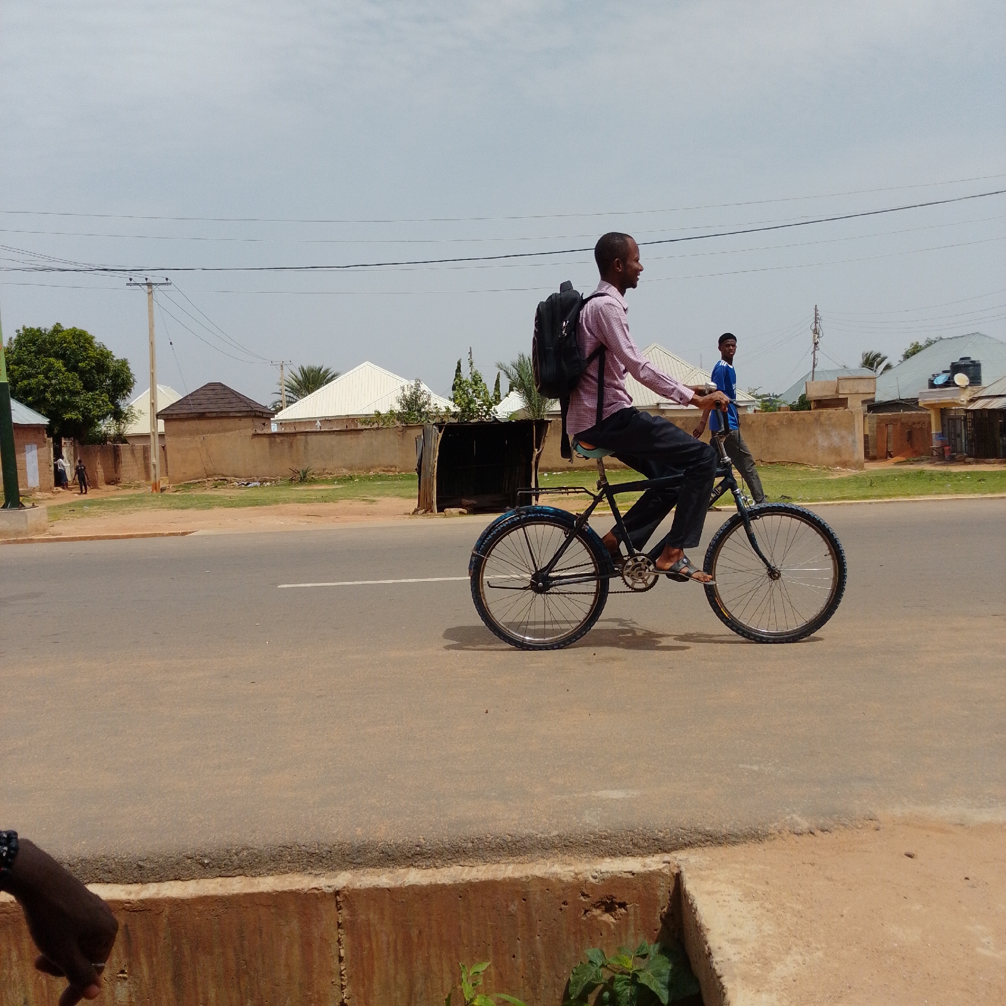 In Gombe, Nigeria, a man wearing a pink checkered shirt, dark pants, and sandals rides a bicycle with a black backpack. The bicycle has a dark frame and blue rims. He is on a paved road with a white line marking the lane. Behind him, another man in a blue shirt and dark pants walks on the side of the road. The background shows buildings with white roofs, a concrete wall, trees, and utility poles with overhead wires under a cloudy sky. The foreground shows a concrete barrier.