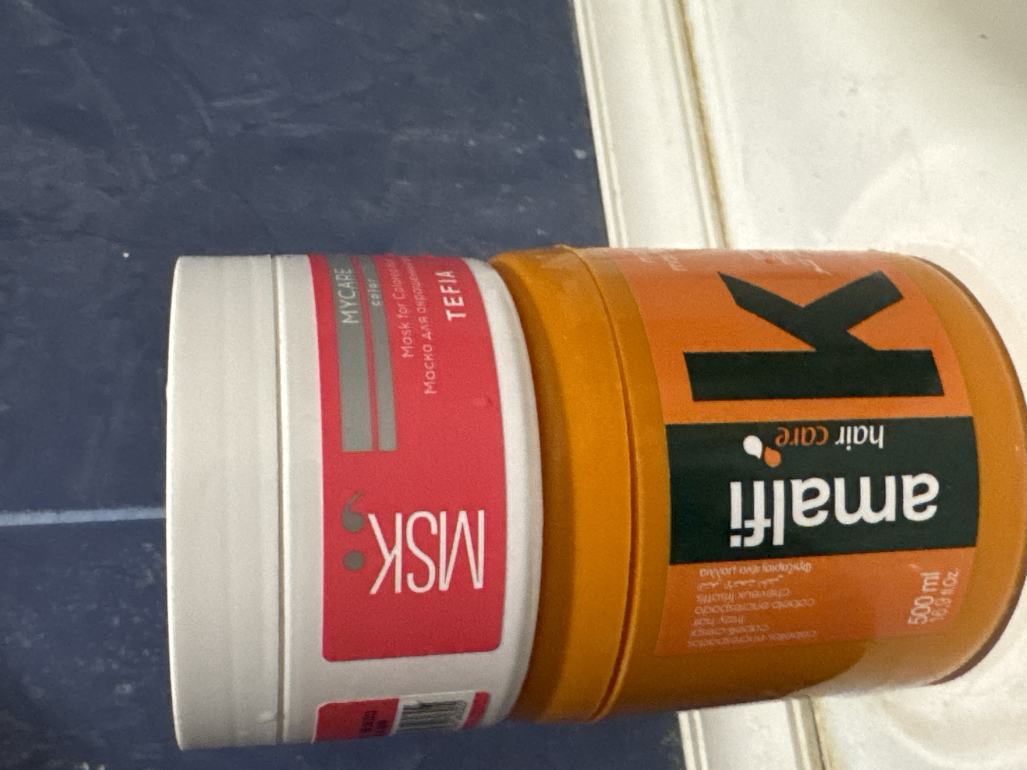 In a brightly lit indoor setting, two hair care products are stacked on a white surface, possibly a bathroom counter. The top container is white with a red label that reads "MSK" and "MYCARE color Mask for Colored Hair". The bottom container is orange and features a prominent black label with white text that says "amalfi hair care" and a large "K". The background appears to be a blue tiled wall. There are no people or animals present, and no actions are being taken. The scene is static and focused on the products. The location of Яромінскі сельскі Савет, Belarus, is not evident from the visual information provided in the image.