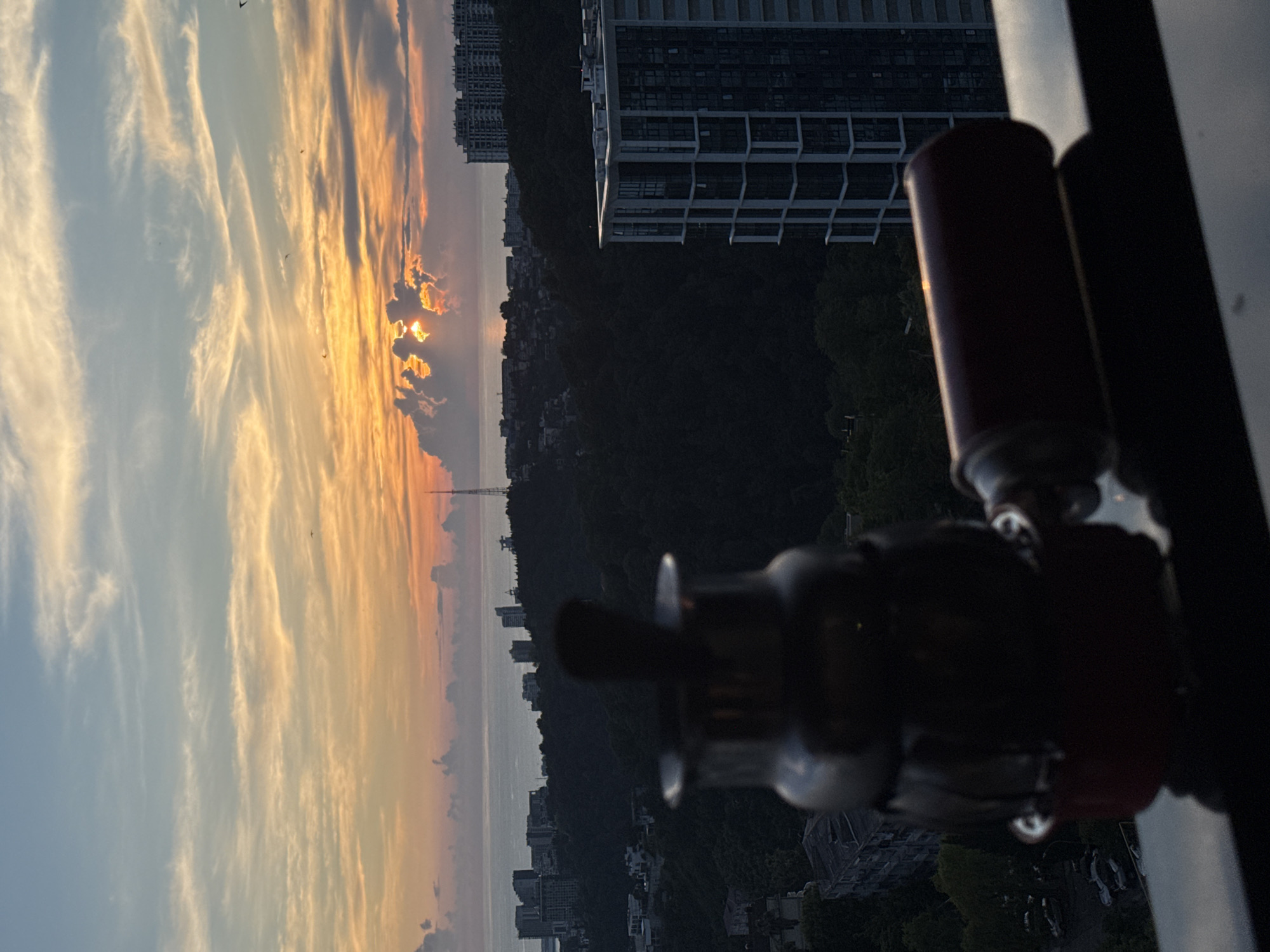 The image captures a sunset over the city of Sochi, Russia. In the foreground, a blurred metal object, possibly a coffee maker or a samovar, sits on a dark surface, likely a balcony railing. To the right of this object is a cylindrical, dark red item, also out of focus. The background reveals a city skyline with buildings of varying heights, nestled amongst dark, tree-covered hills. The sea is visible in the distance, meeting the horizon under a sky filled with dramatic clouds tinged with orange and yellow from the setting sun. The lighting suggests it is late afternoon or early evening. No people are visible in the frame.