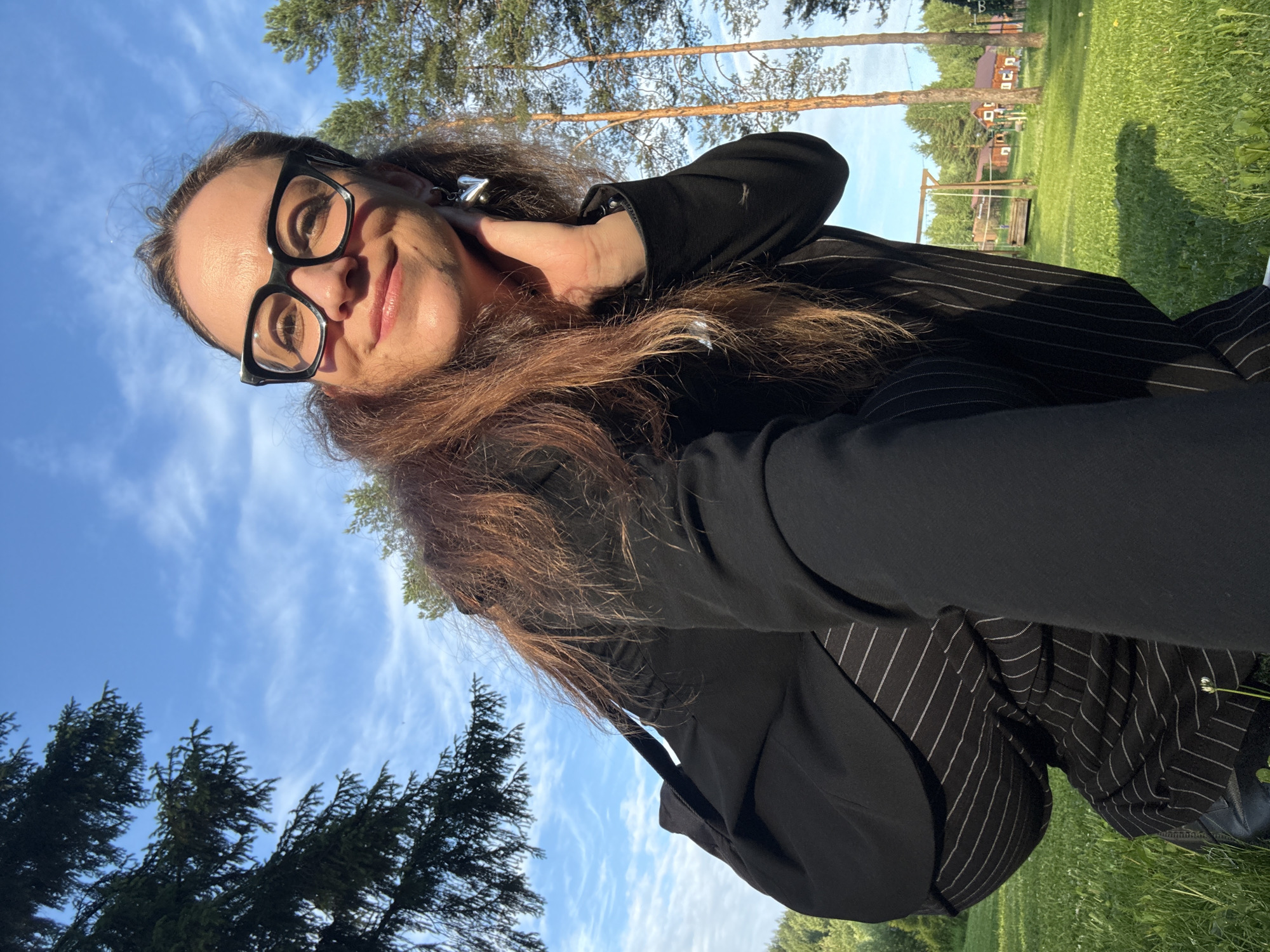 In this selfie, a woman with long, brown hair and glasses looks at the camera with a smile. She is wearing a black jacket over a pinstriped outfit. Her hand is raised to her neck, and she appears to be in an outdoor setting. The background shows a clear blue sky with some wispy clouds, pine trees, and a grassy area with a wooden swing set and houses in the distance, suggesting a rural or suburban location. The lighting indicates it is daytime, likely late afternoon due to the warm sunlight casting shadows. The woman's expression suggests happiness and relaxation. The image was taken in Votkinsk, Russia.
