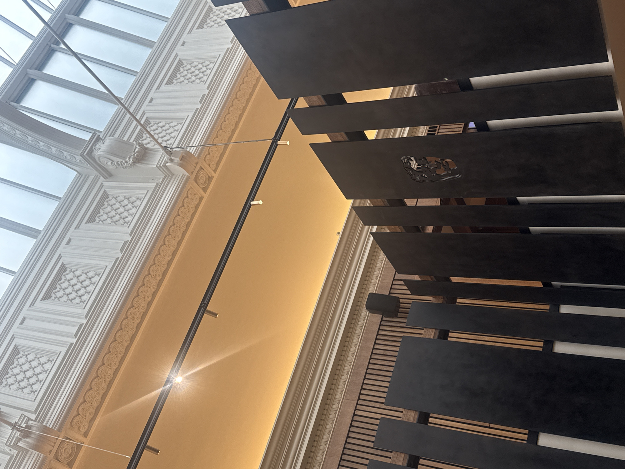 The image displays an architectural interior, looking upwards at a dynamic, oblique angle. On the left, a large, multi-paneled glass skylight allows diffused natural light to enter the space, suggesting daytime. Below the skylight, an ornate white plaster ceiling or wall section is visible, adorned with intricate classical detailing. This includes square coffers featuring a diagonal grid pattern, decorative cornices with continuous leaf or heart-shaped motifs, and a sculpted element resembling a wreath or scrollwork.
Traversing the middle ground is a dark, linear lighting fixture suspended by thin wires, equipped with several small, bright spotlights that cast a starburst effect. This fixture illuminates a warm, golden-hued wall or ceiling section that extends across the upper-middle of the frame.
To the right, a contemporary architectural feature is prominent: a series of dark, horizontal rectangular slats. These slats are backed by parallel wooden strips, creating a textured, modern screen or wall treatment. One of the dark slats distinctly features an intricate, cut-out design, possibly a stylized animal head or a heraldic crest. The overall composition highlights a striking contrast between the historical, classical plasterwork and the sleek, modern elements.
No people are present in the image, nor is there any visible text. The setting appears to be a grand hall, atrium, or a similarly significant interior space, blending traditional grandeur with modern design, typical of a renovated or architecturally rich building in London, United Kingdom.