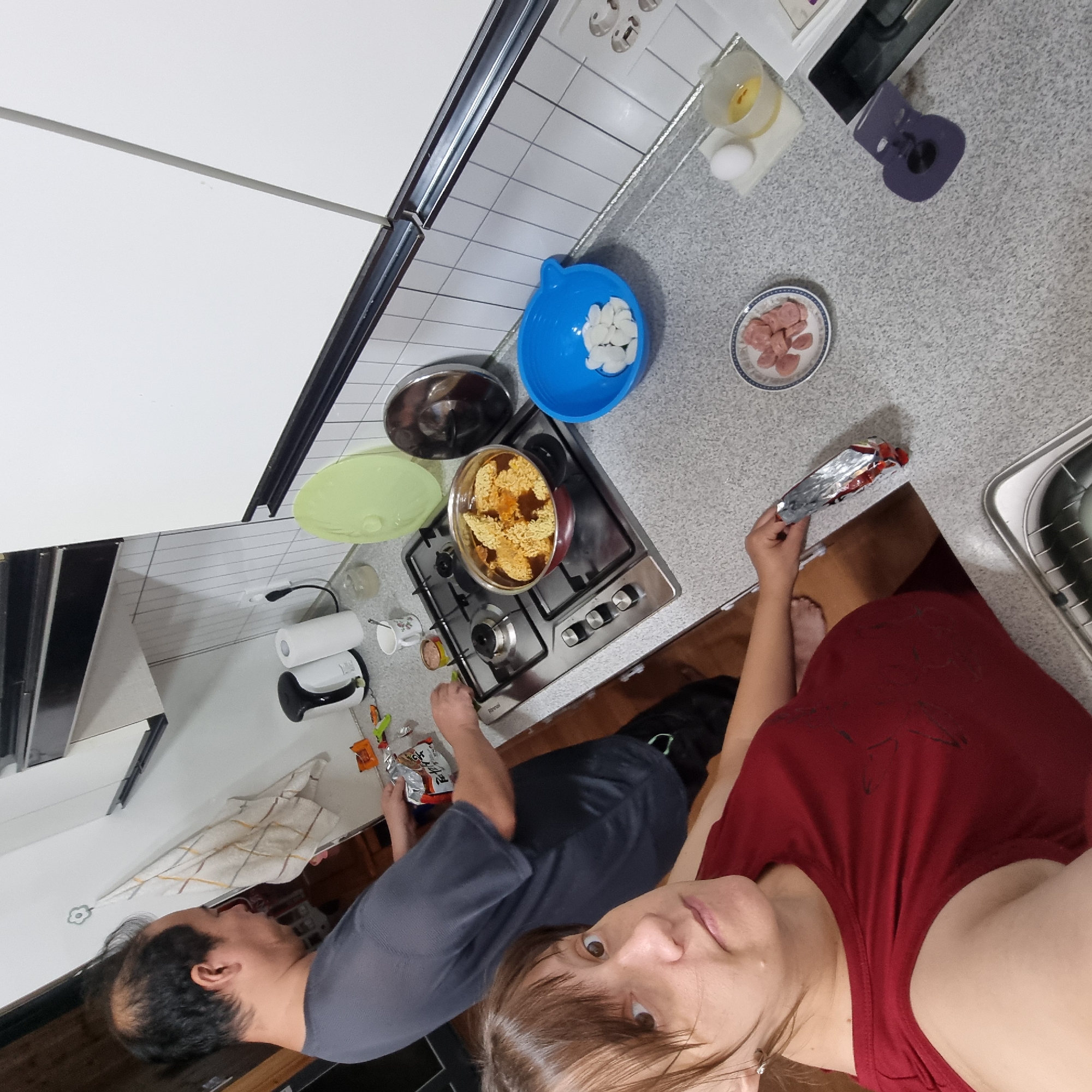 In a domestic kitchen in Dunnae-myeon, South Korea, a man and a woman are preparing food. The man, wearing a grey t-shirt, stands near a gas stove with a pot of ramen noodles and broth cooking. He is holding a package of instant ramen seasoning. The woman, wearing a red tank top, is in the foreground, looking towards the camera. The kitchen counter has several items including a blue bowl with white rice cakes, a small plate with sliced sausages, an egg, and a measuring cup with egg yolk. A roll of paper towels and a kettle are also visible. The overall scene suggests a casual meal preparation.