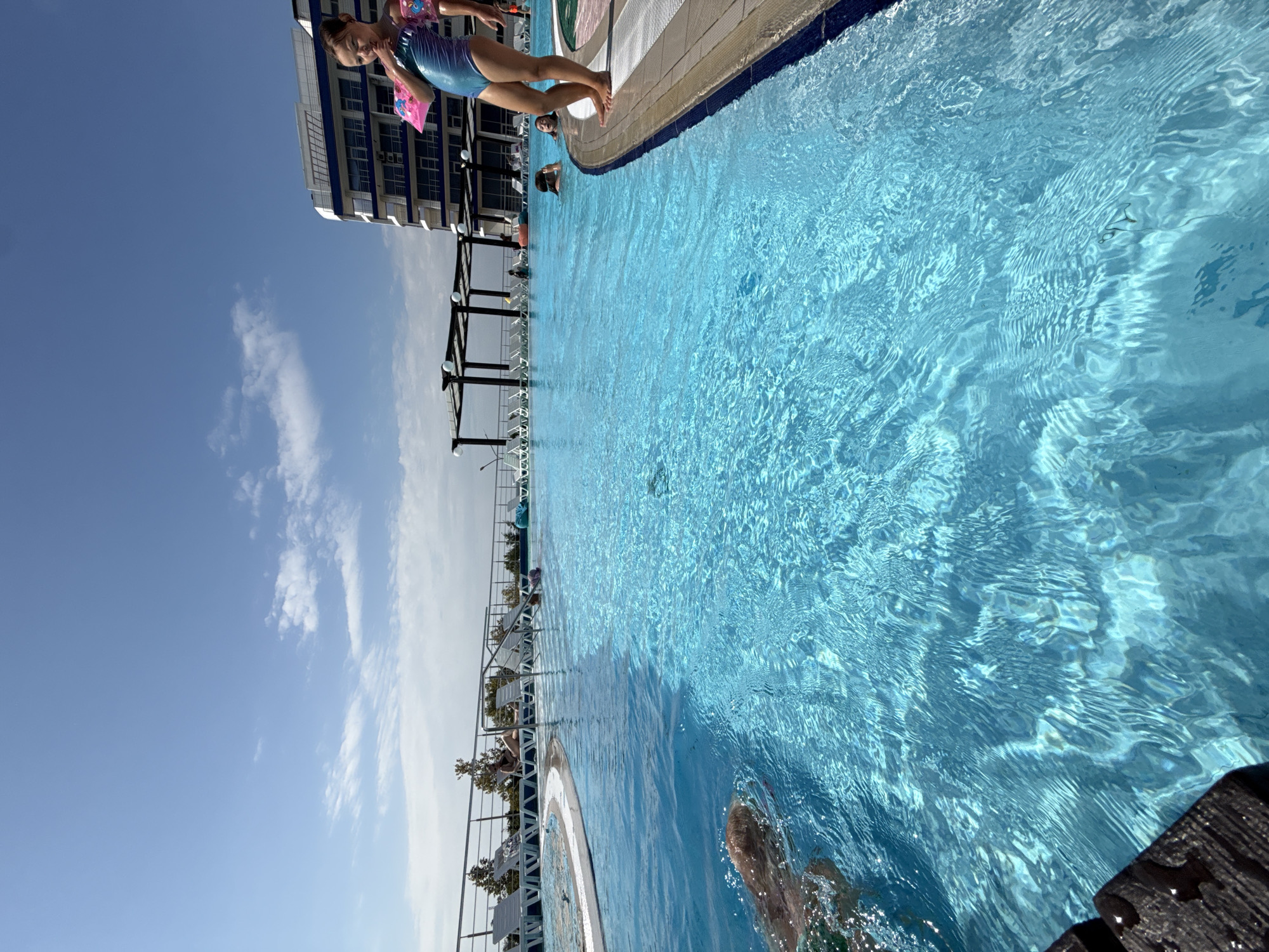This is a ground-level, wide-angle, eye-level photograph of an outdoor swimming pool on a sunny day. The pool's clear blue water fills most of the frame, with ripples and reflections creating a dynamic surface. In the foreground, the dark, wet edge of the pool's deck is visible on the left. A person's head and shoulders are submerged in the water on the left side of the frame, appearing to swim towards the camera.
In the middle ground, several people are in the pool. Two children are visible in the water, one wearing pink arm floaties. Further back, a person is lying on a lounge chair by the pool. The pool area is lined with lounge chairs and umbrellas.
In the background, a modern, multi-story building with blue and white accents stands behind the pool area. The sky above is a clear blue with scattered white clouds. The overall scene depicts a resort or hotel pool area, likely in Sevastopol, Ukraine, given the context.