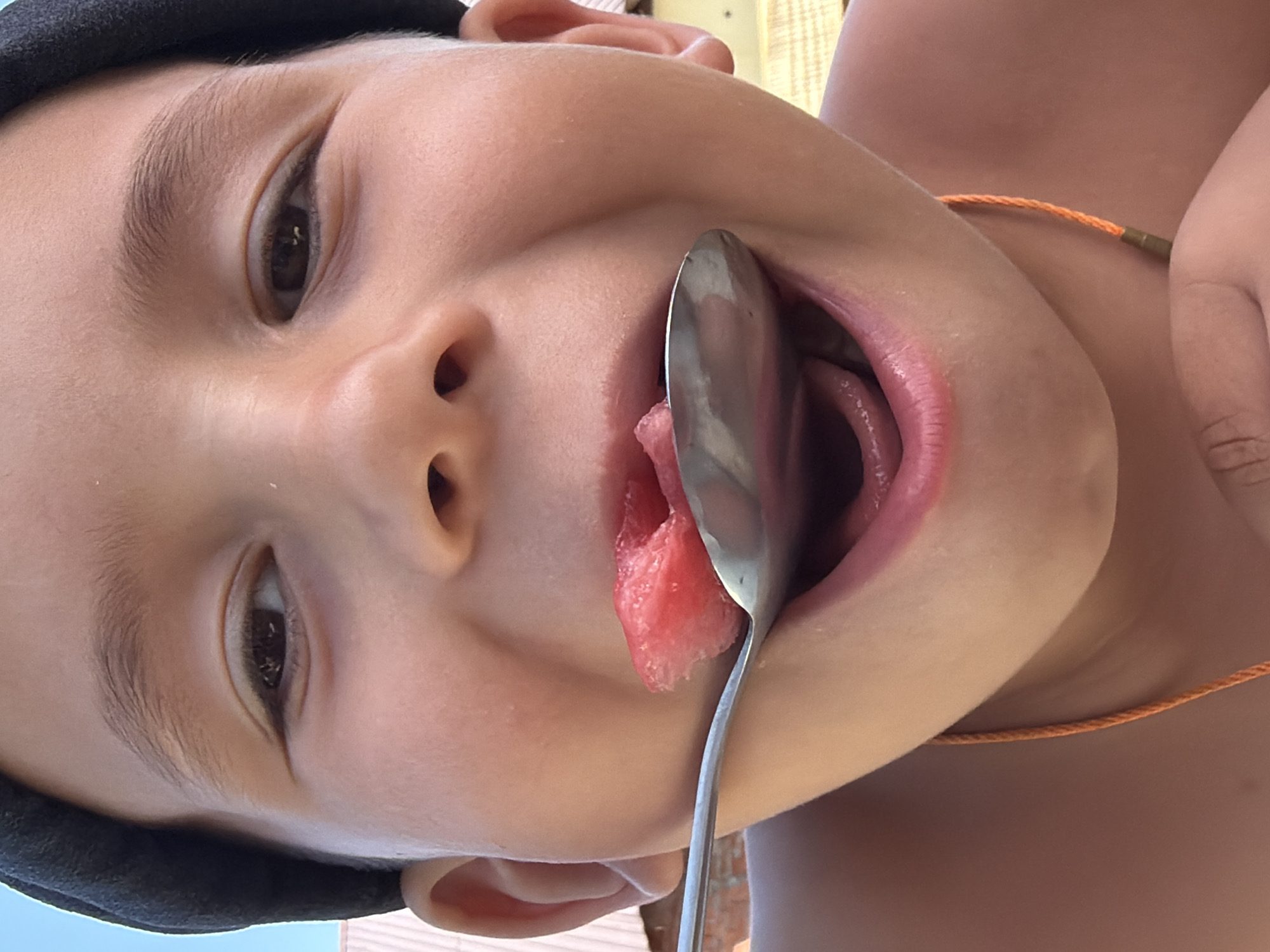 A close-up, low-angle shot shows a young boy's face as he eats watermelon from a spoon. The boy is wearing a dark beanie and an orange string necklace. His mouth is open, revealing his tongue and teeth, and a piece of watermelon is on the spoon, about to enter his mouth. The background is blurred, but appears to be an outdoor setting with some structures visible. The image was taken in Chaikovsky, Russia.