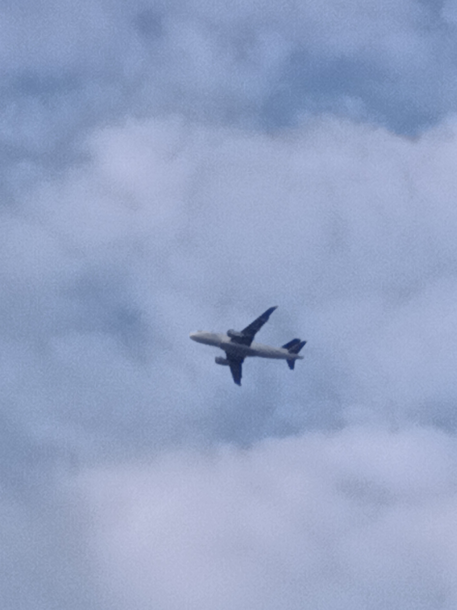 A commercial airplane is captured in mid-flight against a backdrop of a cloudy sky. The aircraft is light-colored with visible wings, engines, and tail. The sky is filled with various shades of blue and white clouds, creating a diffused lighting condition. The image is slightly grainy, suggesting it may have been taken with a low-resolution camera or under challenging lighting conditions. There is no indication of the location being Prague, Czechia.