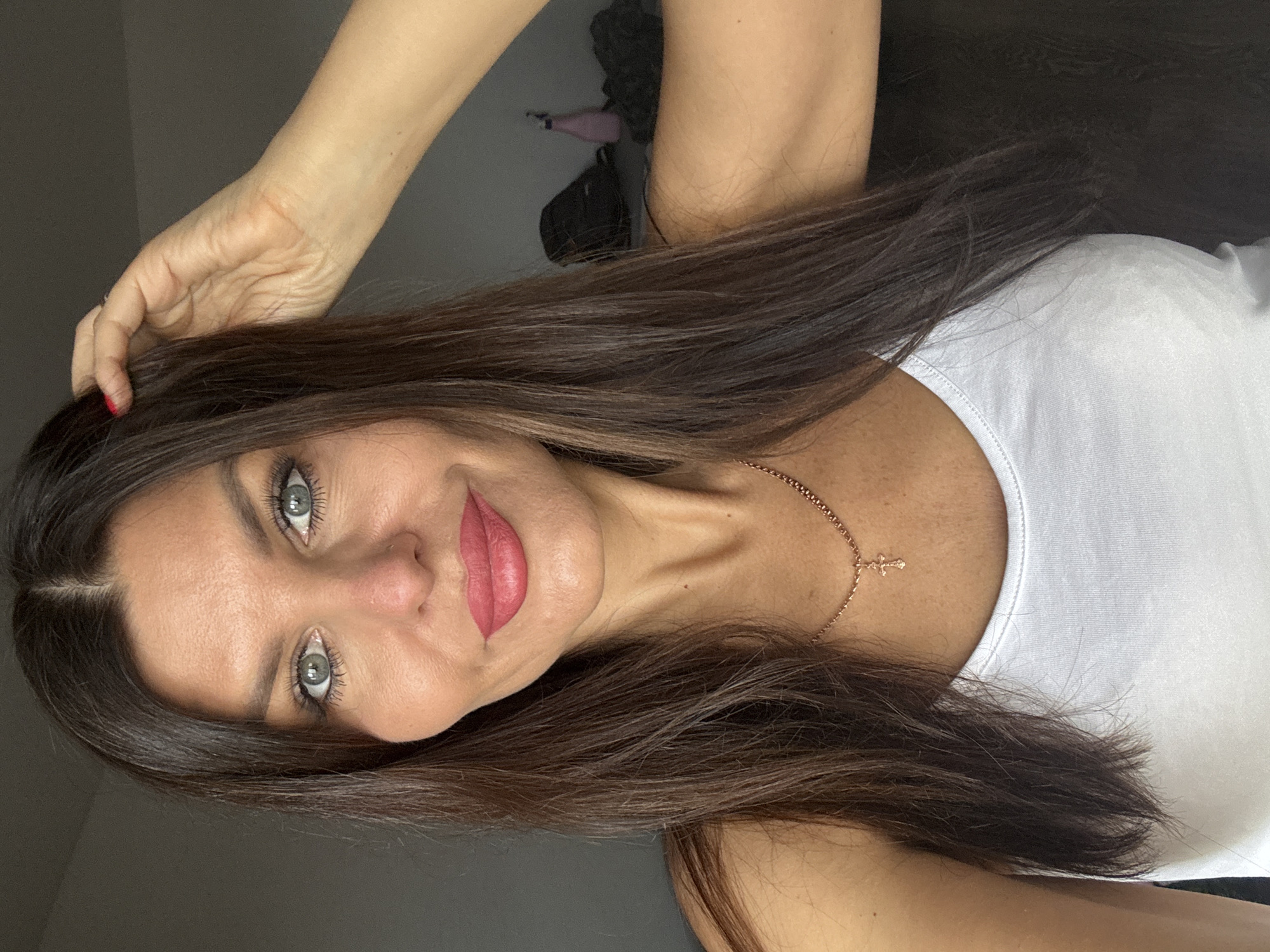 A woman with dark brown hair and blue eyes, wearing a white tank top and a rose gold necklace with a cross pendant, poses for a selfie. She is touching her hair with her right hand. Her makeup includes bold pink lipstick and mascara. The background is blurred and appears to be an indoor setting, possibly a bedroom, with a dark wall and some objects, including a pink bottle and a dark bag, visible on a surface. The image does not contain any information indicating the location within Chaikovsky, Russia.