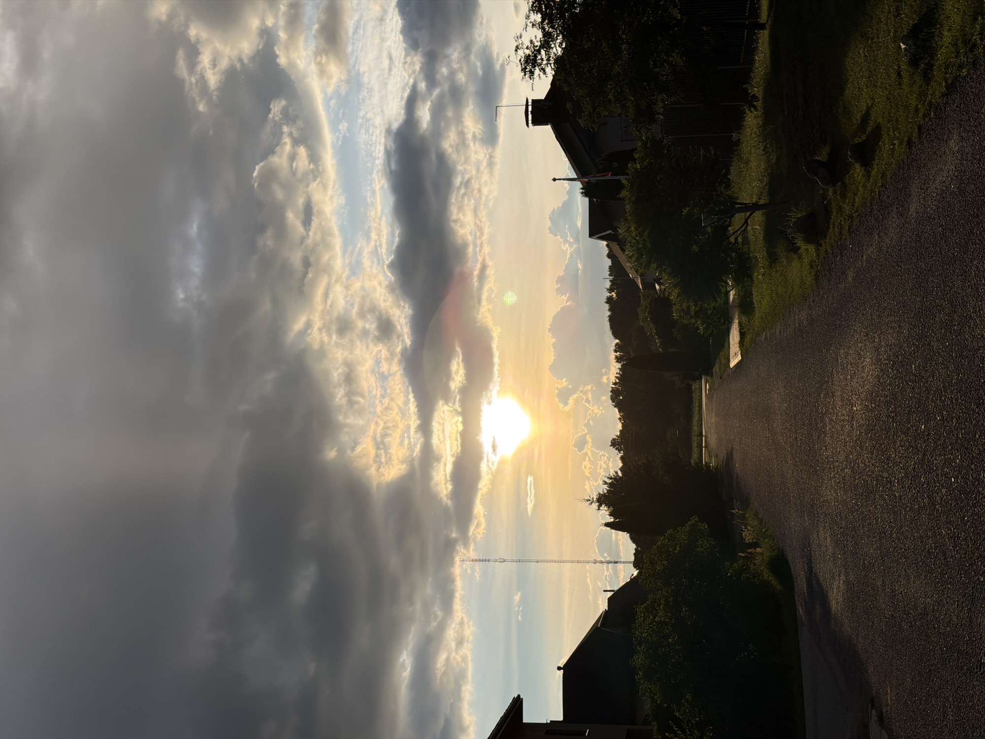The image depicts a residential street in Ulbroka, Latvia, at sunset. The sky is filled with dramatic, heavy clouds, with patches of bright sunlight breaking through. The sun itself is low on the horizon, casting a warm, golden glow on the scene and creating lens flare. On the left side of the image, the silhouette of a house with a dark roof is visible, partially obscured by lush green bushes. A tall, slender radio tower stands in the distance, between the houses. On the right side of the street, another house with a brick chimney and a flag pole is visible, also silhouetted against the sky. A dense line of trees forms a dark backdrop beyond the houses. The street itself is paved with asphalt and appears to be gently sloping upwards. The surrounding environment includes well-maintained lawns and grass verges. The overall atmosphere is one of a quiet, suburban evening as day transitions into night.