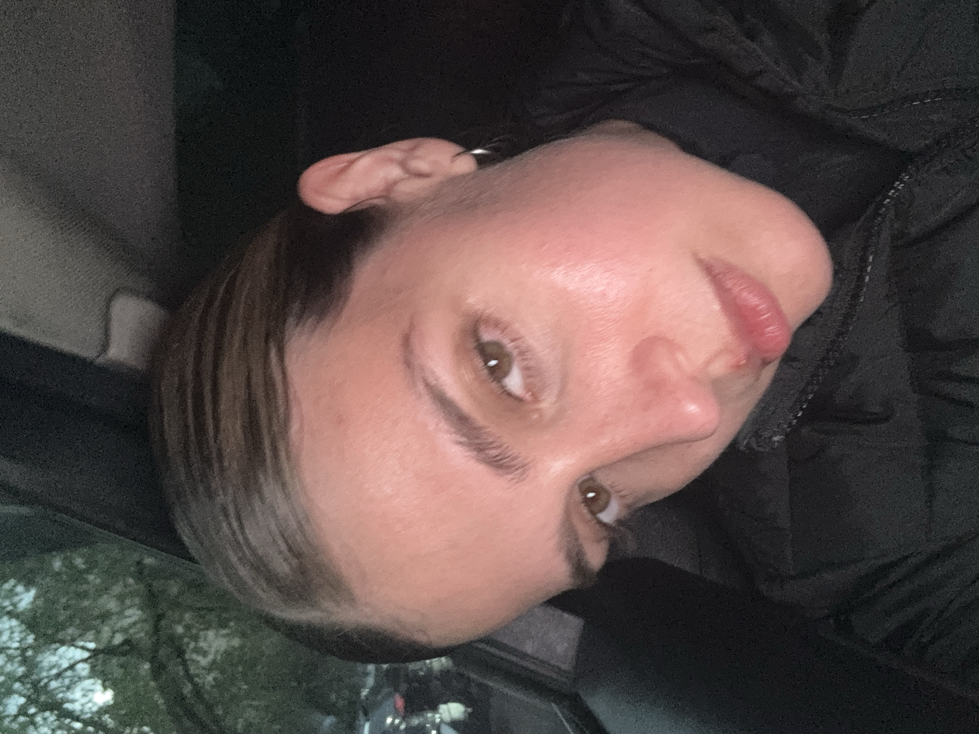 The image presents a close-up portrait of a person, likely a woman, with light skin, brown eyes, and dark brown hair that appears styled back, possibly with a slicked-back effect showing distinct strands. The image is oriented with the person's head tilted to the right, implying it might be a selfie or a photo taken in a landscape orientation and then rotated.
The subject is wearing a black puffy jacket, which is visible on the right side of the frame and around their neck. A small silver hoop earring can be seen on the upper part of their left ear. Their lips are slightly parted, and they have a neutral to slightly pensive expression, with their gaze directed off to the side.
The setting appears to be inside a vehicle or a dimly lit interior. On the left side of the frame, through what seems to be a car window, green foliage and tree branches are visible, indicating an outdoor background, possibly during overcast daylight or twilight hours. The interior parts of the scene are dark, creating contrast with the relatively well-lit face of the subject. No visible text is present in the image.