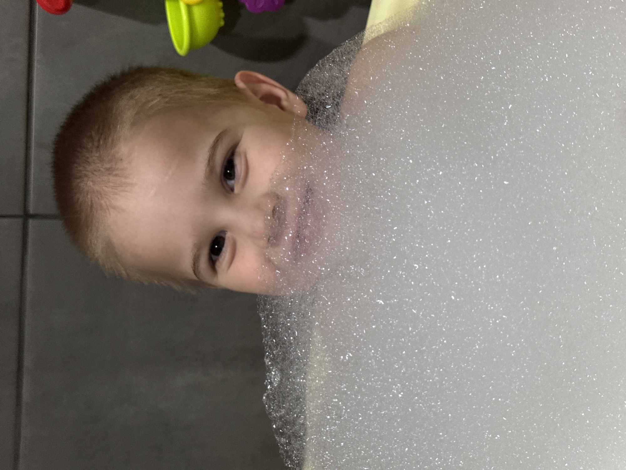 A young child is partially submerged in a bathtub filled with white soap bubbles. The child is looking directly at the camera with a slight smile. The child has short blond hair and fair skin. In the background, out of focus, there are colorful bath toys. The photograph was taken indoors, likely in a private bathroom with grey tiles on the wall. There is no information provided in the image to confirm the location is in the city of Chaikovsky, Russia.