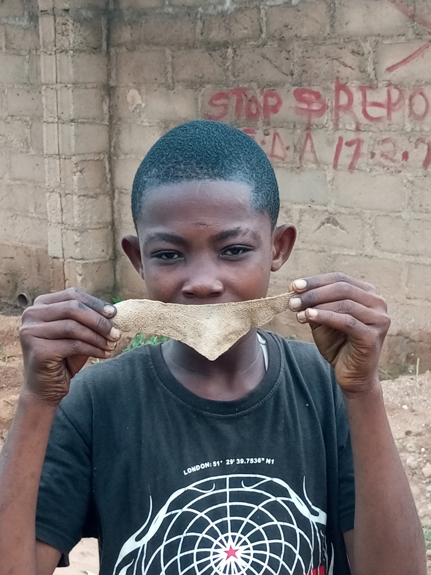 A young Black male, likely a child or adolescent, is pictured in an outdoor, possibly urban environment, with a rough brick wall as the backdrop. The wall has red spray paint graffiti on it, partially visible reading "STOP REPO" and "17.2.7". The subject is wearing a black t-shirt with a white circular design on it. He is holding a piece of light brown, textured material, possibly a strip of leather or cloth, in front of his mouth with both hands, obscuring his lower face. His expression is neutral or slightly inquisitive as he looks directly at the camera. The ground appears to be dusty or sandy with some scattered stones. The lighting suggests it is daytime. The location is identified as Tempure, Nigeria.