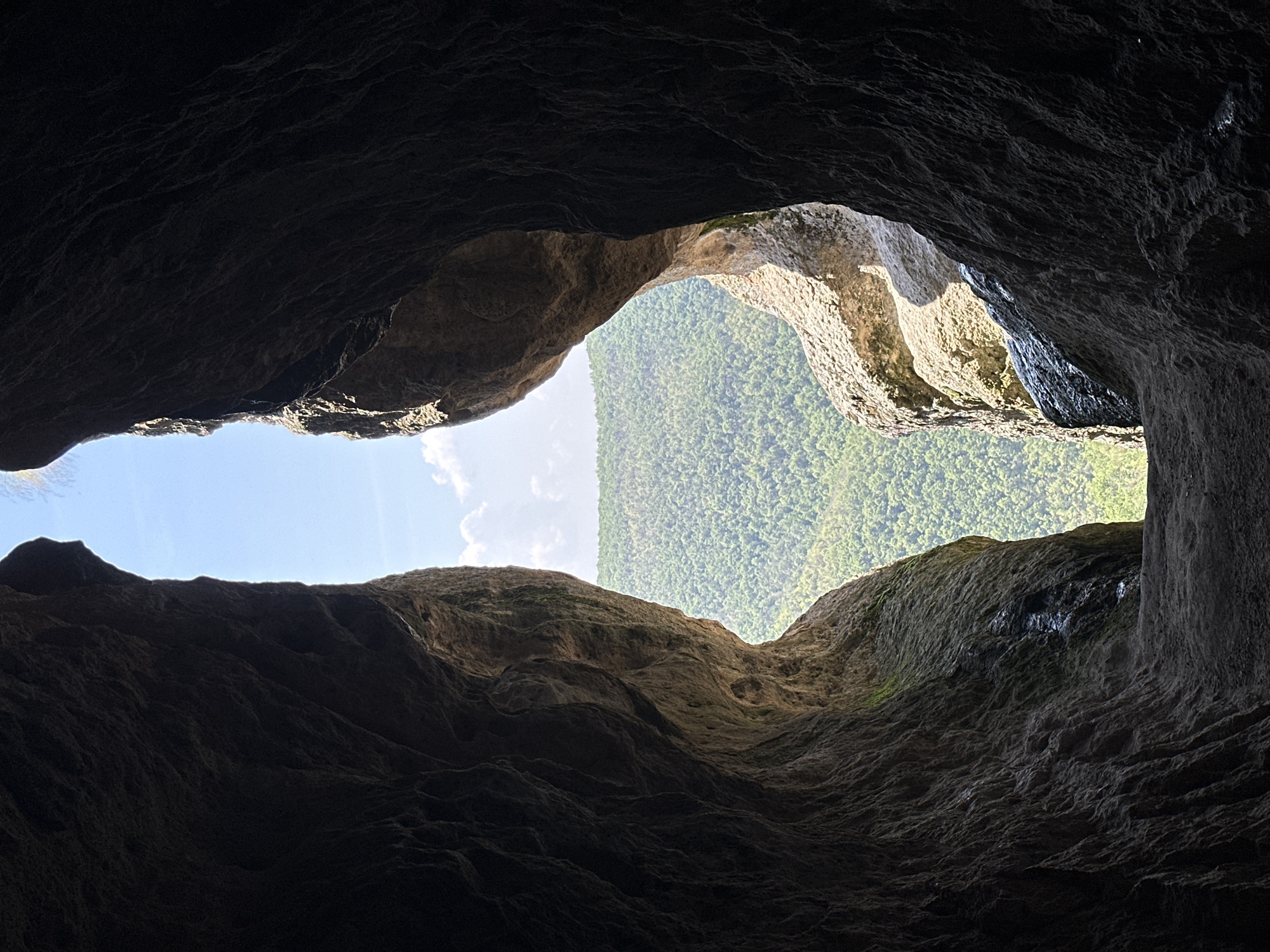 The image displays a forensic view from within a dark, natural rock cave opening, located in Ilinitsa, Bulgaria. The foreground is dominated by the interior of the cave, characterized by dark, heavily textured, and undulating rock walls that frame the central exterior view. These cave walls show varied shades of dark grey and brown, with some sections on the right exhibiting subtle indications of moisture or moss. The opening itself is an irregularly shaped, vertical aperture, wider at the top and narrower towards the bottom, forming a natural archway.
Through this opening, a brightly illuminated outdoor scene is visible. The upper portion reveals a clear blue sky with several distinct white cumulus clouds. Beneath the sky, the midground and background consist of a densely forested landscape, showcasing vibrant green deciduous or coniferous trees covering undulating terrain, likely hills or mountains, stretching into the distance. Direct sunlight strongly illuminates the exterior landscape and the outer rock formations on the right side of the opening, creating a significant contrast with the deeply shadowed interior of the cave. The ground surface directly at the threshold of the cave opening appears dark, rocky, and uneven. There are no identifiable people, animals, or man-made objects within the frame.