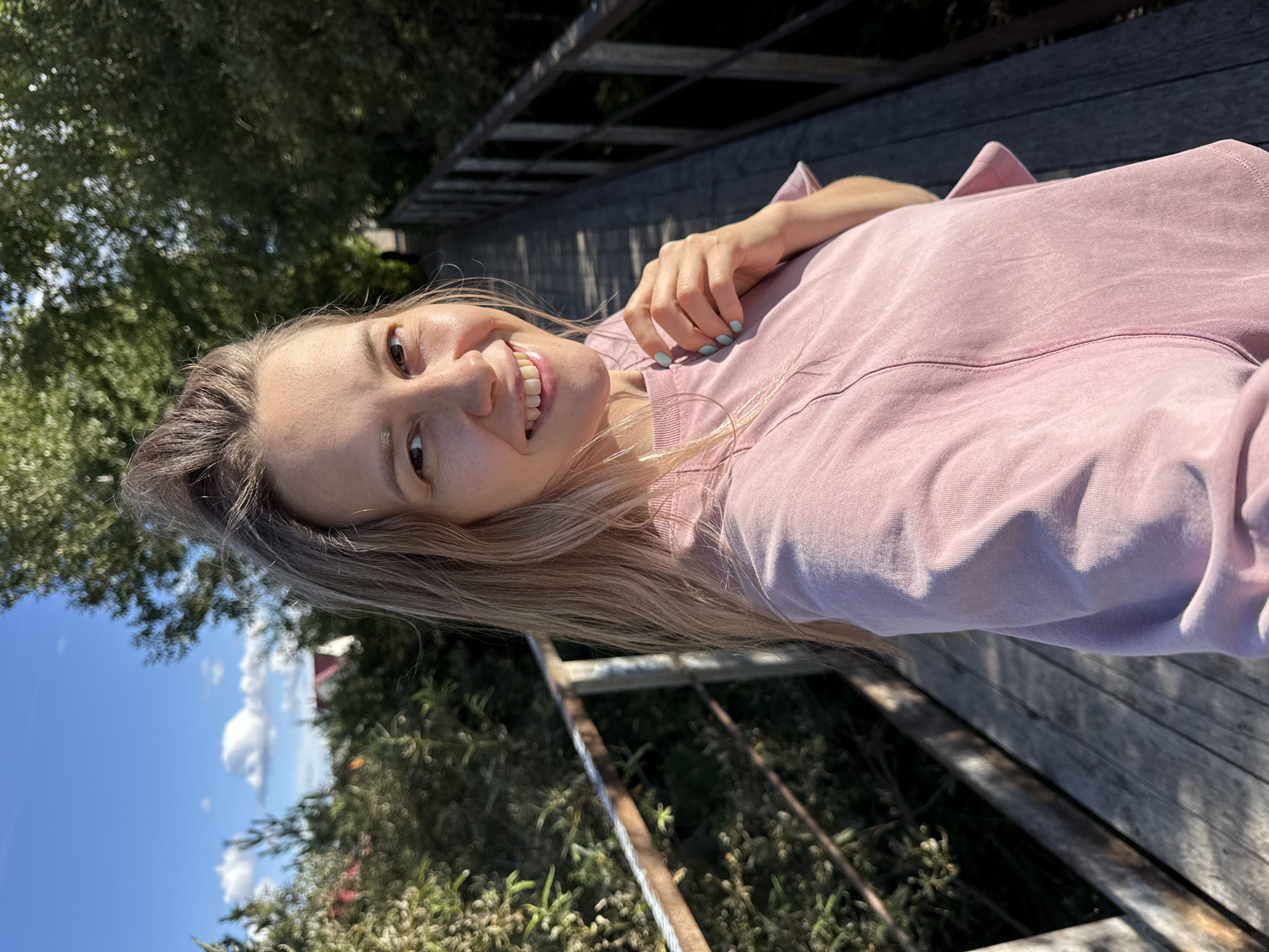 A woman with blonde hair tied up in a bun is standing on a wooden bridge, taking a selfie. She is wearing a pink t-shirt. The bridge is flanked by trees and foliage. In the background, there are houses and a clear blue sky with some clouds. The woman is smiling and looking at the camera. Her fingernails are painted a light blue color.