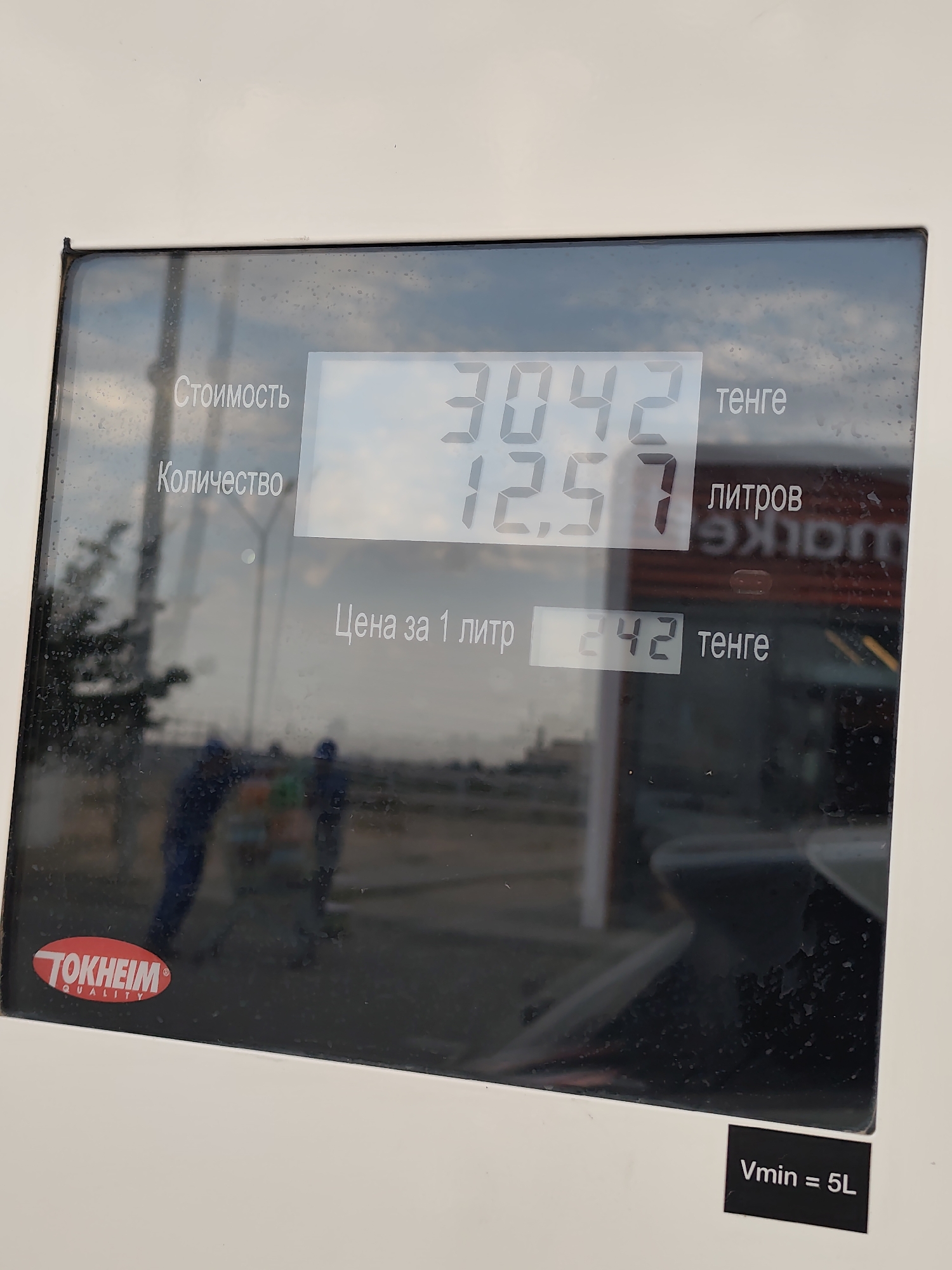 The image shows a close-up of a digital display on a fuel pump. The display shows three lines of information. The top line reads "Стоимость" (Cost) and displays "3042" in large digits, followed by "тенге" (tenge), the currency of Kazakhstan. The second line reads "Количество" (Quantity) and displays "12.57" in large digits, followed by "литров" (liters) and a reflection of the word "TOKHEIM". The third line reads "Цена за 1 литр" (Price per 1 liter) and displays "242" in large digits, followed by "тенге". A red and white "TOKHEIM QUALITY" logo is visible in the bottom left corner of the display. The background of the display shows a reflection of a cloudy sky and a street scene with two individuals wearing blue uniforms and two small carts. A sticker on the bottom right corner reads "Vmin = 5L". The context provided places this scene in Ömirzaq, Kazakhstan, at a fuel station.