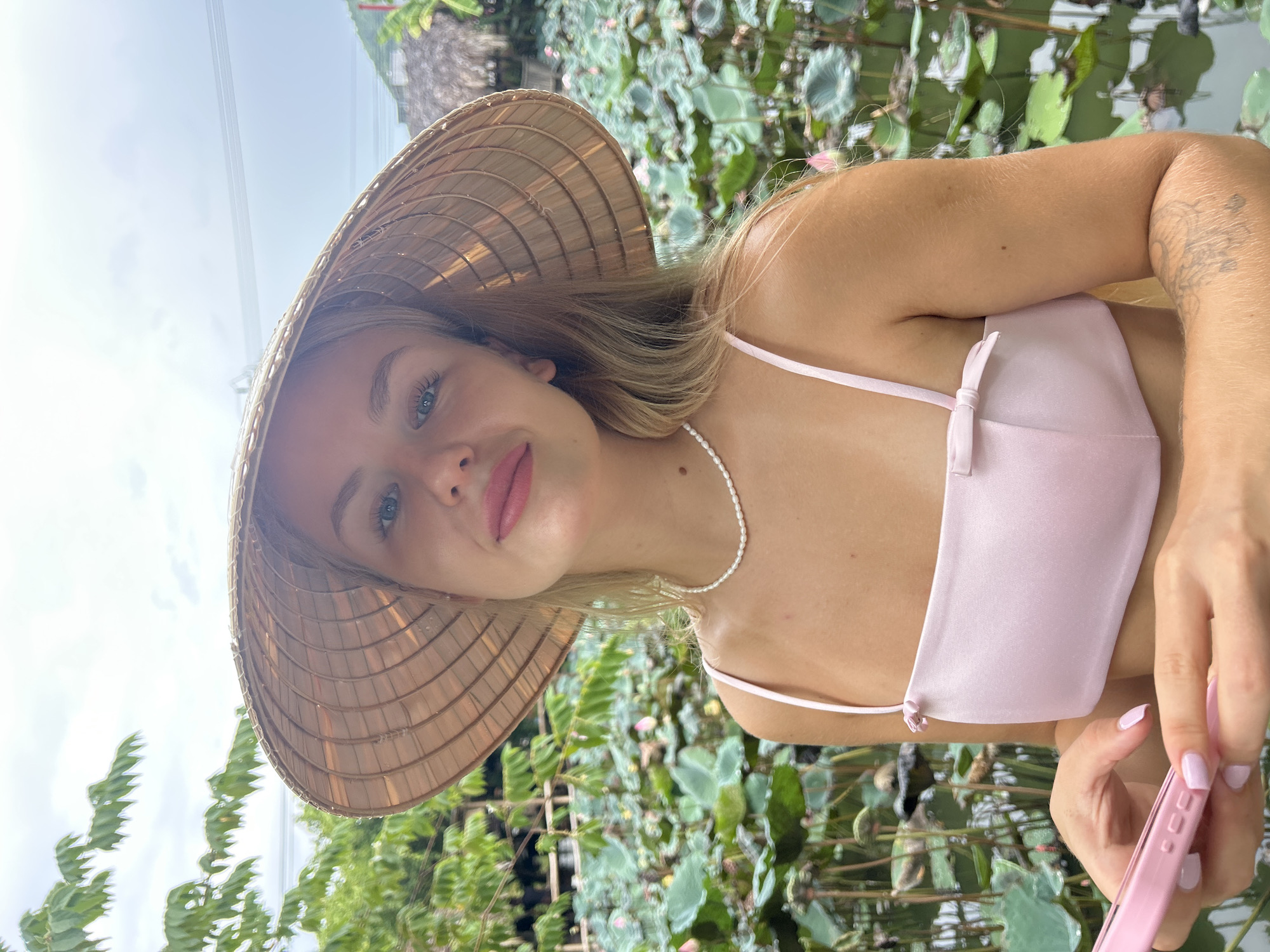 A young Caucasian woman wearing a conical hat and a pink top with a bow is holding a pink phone and looking at the camera. She has blonde hair, blue eyes, and is wearing a pearl necklace. She is standing in front of a pond filled with lotus flowers. The background shows greenery and a cloudy sky. The scene appears to be in a natural setting, possibly a botanical garden or a rural area in Nha Trang, Vietnam.