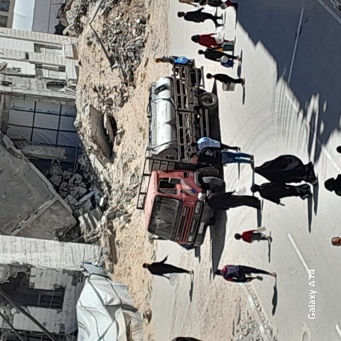 This image depicts a street scene in Khan Yunis, Palestinian Territory, marked by extensive urban destruction. In the background, multiple concrete buildings are severely damaged or partially collapsed, with large quantities of rubble, rebar, and sand debris accumulated at their bases. A white, possibly canvas, tent structure is visible on the far left.
In the midground, a red, forward-control truck with a silver cylindrical water tank mounted on its rear flatbed is positioned on a paved road. Approximately 15 to 20 individuals are present across the frame, engaged in various movements. People are walking in multiple directions, many carrying white plastic containers, bags, or other items. Notable individuals include a person in dark clothing walking left and carrying two white containers; a small child in a red shirt walking right, holding a white object; two figures, likely women, in black abayas or niqabs walking away from the viewer in the foreground; and a group of individuals moving from right to left across the midground behind the truck, several of whom are carrying bags or containers. One person is seated on the rear of the water tanker truck. The scene is illuminated by strong daylight, casting prominent shadows on the paved road.