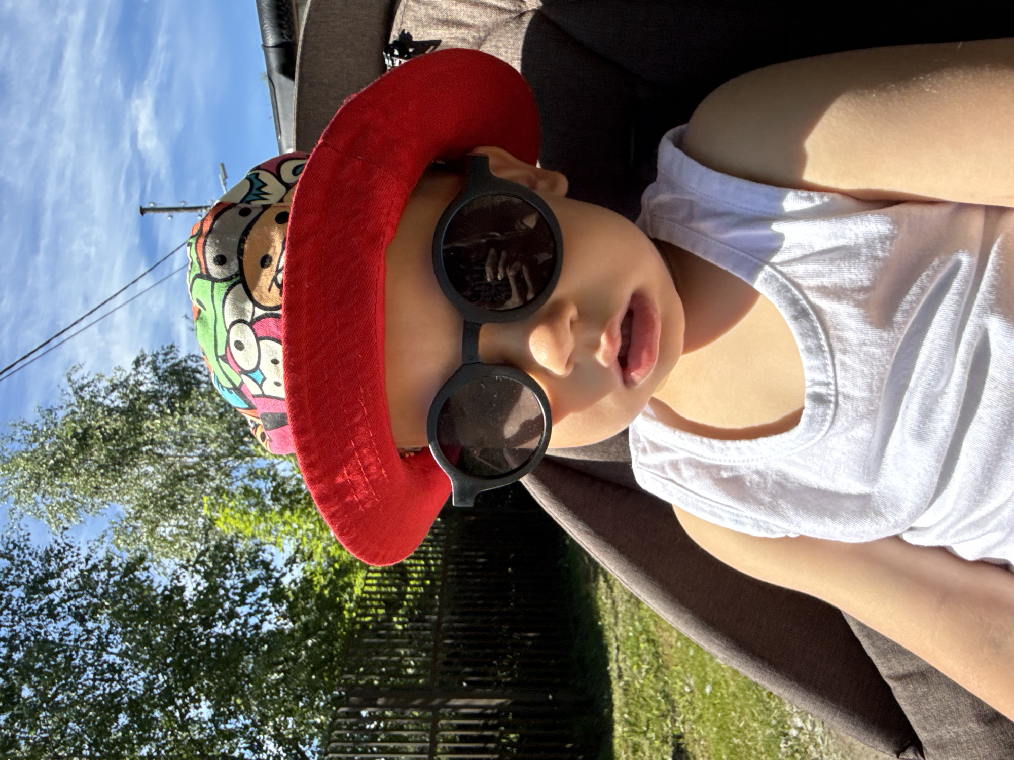 A young child, likely a toddler, sits in a stroller outdoors in Rusanovo, Russia. The child wears a red wide-brimmed hat with a colorful cartoon pattern on top and dark, round sunglasses. They are dressed in a white sleeveless shirt. The child's face is partially obscured by the sunglasses and hat. Behind the child, a dark brown stroller seat is visible. The background shows a wooden fence, green grass, trees, and a bright blue sky with scattered clouds, suggesting a sunny day. The image is taken from a slightly low angle, looking up at the child.