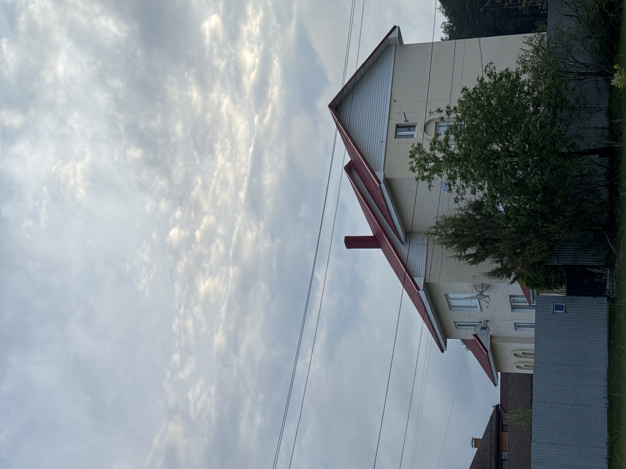 The image shows a multi-story building in Chaikovsky, Russia. The building has a white facade and a red roof. A tree partially obscures the view of the building. Overhead power lines run across the sky, which is filled with white and gray clouds. Another building is partially visible in the foreground. A corrugated metal fence is also visible.