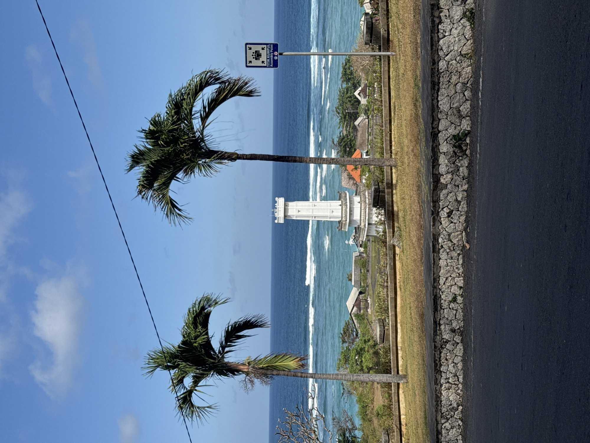In Kuta Selatan, Indonesia, this image captures a bright, sunny day with a clear blue sky and scattered clouds. A dark asphalt road occupies the foreground, bordered by a low, rough-hewn stone wall. Beyond the wall, a strip of dry grass slopes upwards, leading to a paved walkway. Two palm trees stand prominently on either side of the walkway, their fronds casting shadows.
In the middle ground, a white, multi-tiered tower with a crenellated top stands as a central focal point. The tower overlooks a turquoise ocean with visible white-capped waves breaking offshore. A cluster of small buildings with orange and brown roofs is nestled between the tower and the ocean, suggesting a coastal settlement. To the right of the image, a blue and white sign on a pole reads "TITIK KUMPUL Assembly Point" and features icons of people and arrows. The overall environment depicts a scenic coastal view with a mix of natural and man-made elements.