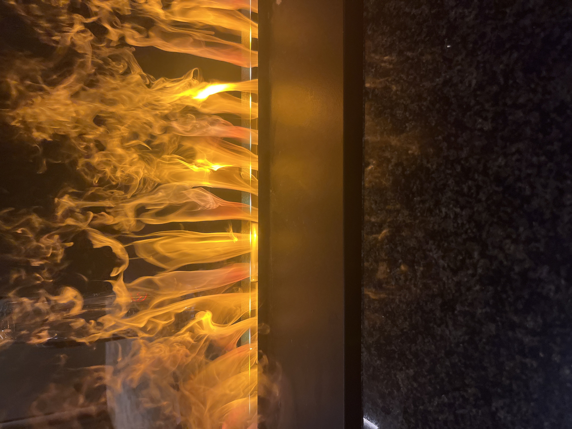 The image presents a striking architectural or artistic installation, likely captured at night or in a dimly lit indoor setting. The primary subject is a vertical display on the left side, featuring highly illuminated, dynamic forms that resemble flowing flames or glowing liquid. These forms are a vibrant orange and yellow, exhibiting intricate, swirling, and wispy patterns that suggest continuous, fluid motion. They appear contained within a transparent column or panel, with distinct horizontal layers contributing to the visual flow.
Adjacent to this luminous display, a dark, solid vertical column or panel serves as a partition, separating the glowing element from the right side of the frame. This column has a smooth, slightly reflective surface. To its right, a dark, textured wall or panel occupies the remainder of the image. This surface appears speckled or granulated, resembling granite or a similar natural stone, providing a stark, dark backdrop to the bright display.
There are no people or discernible activities beyond the implied movement within the illuminated column. The strong contrast between the bright, warm-toned display and the dark surroundings creates an impactful visual, evoking a sense of energy and aesthetic appeal. No visible text is present in the image.